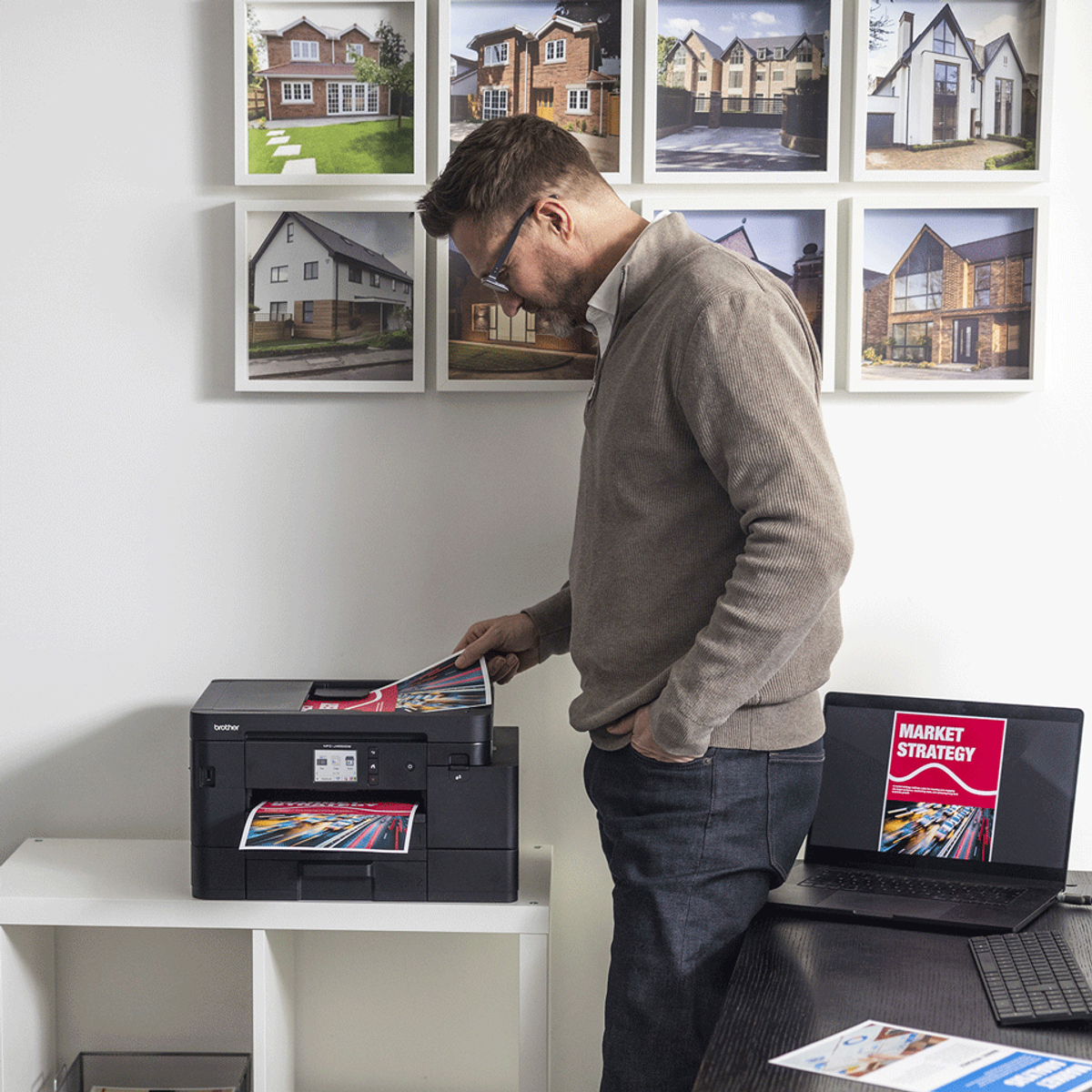 Een man bekijkt een afgedrukt document van een zwarte printer. Een laptop toont 'Marktstrategie'. Foto's van huizen aan de muur.