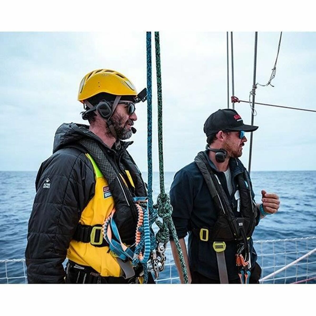 Due uomini su una barca con equipaggiamento di sicurezza. Uno indossa un casco giallo. Guardano il mare. Corde in primo piano.