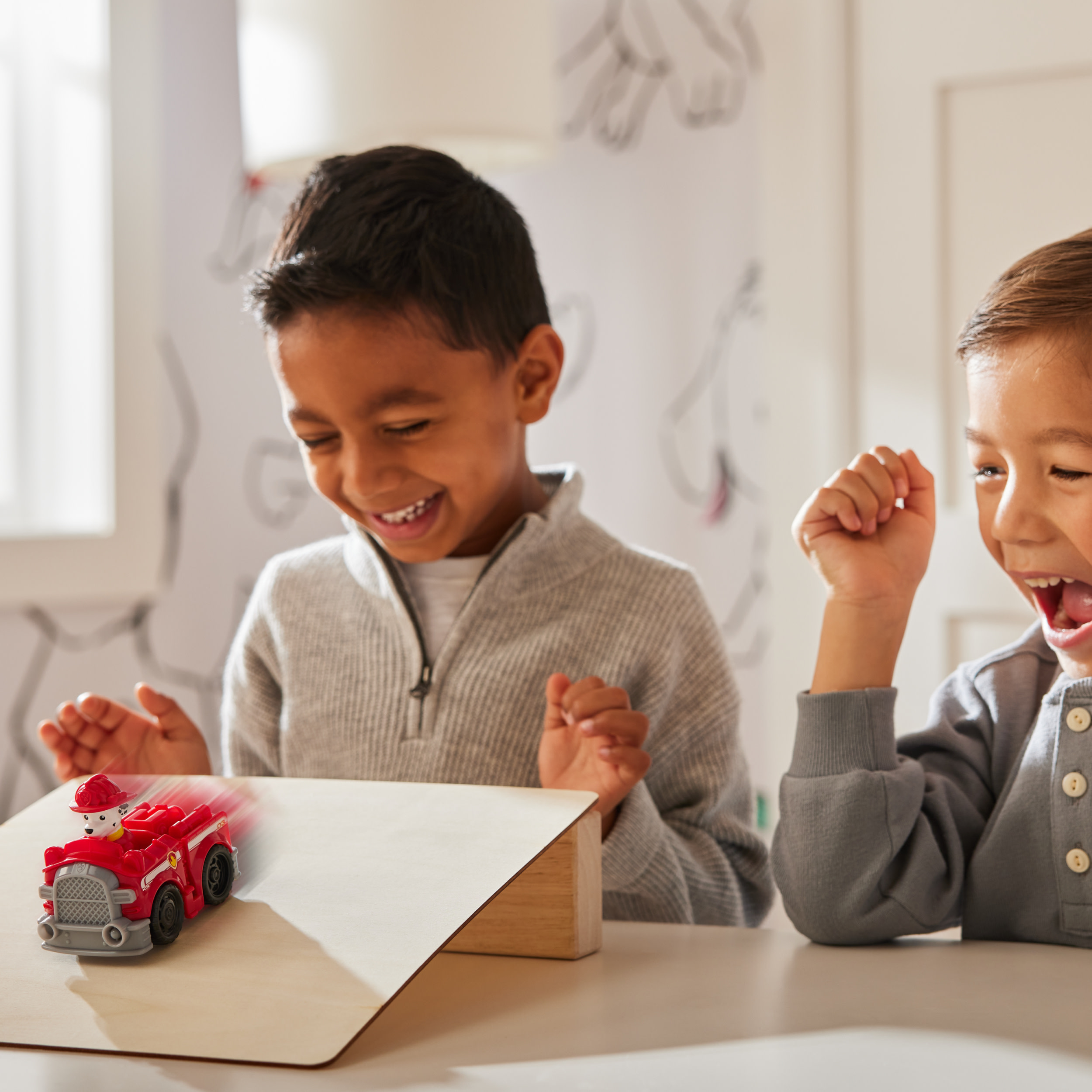 Dwóch chłopców ogląda zabawkową straż pożarną na rampie. Jeden chłopiec się śmieje. Ciężarówka ma rozmycie ruchu.