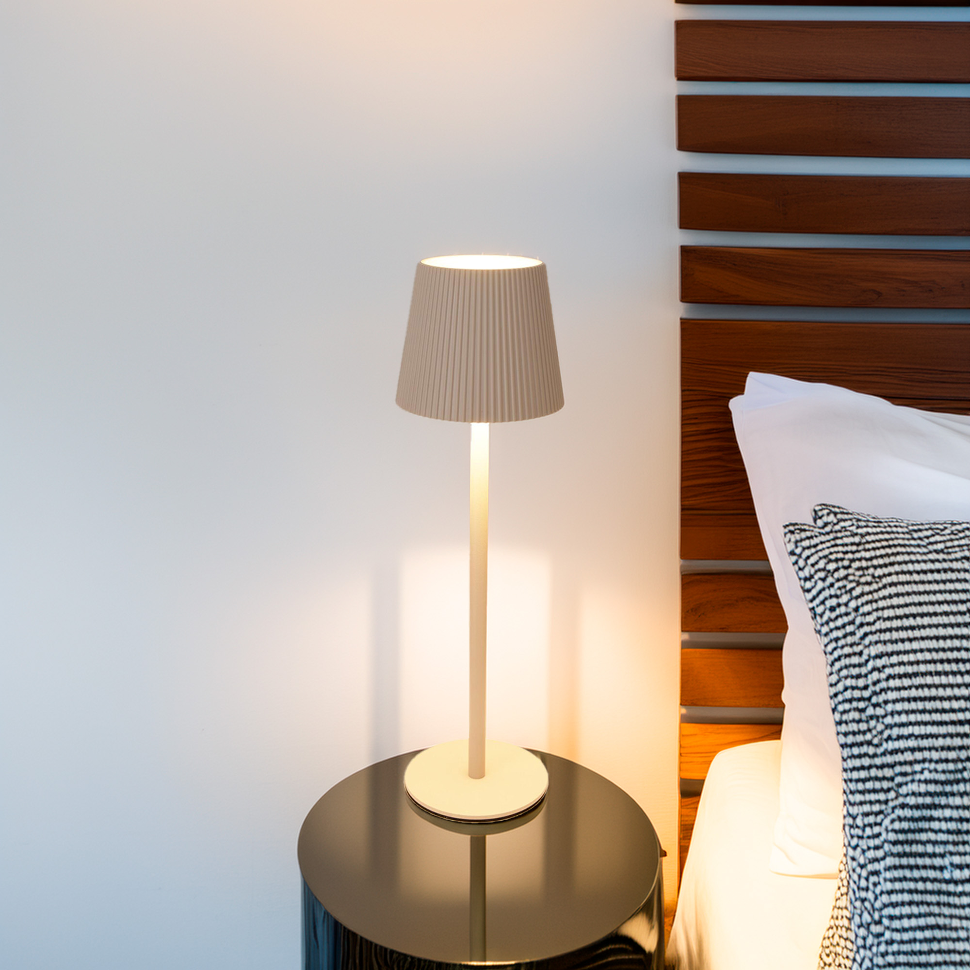 Una pantalla de lámpara beige sobre un soporte blanco se apoya en una mesa negra. A la derecha, una cama con almohada a rayas.
