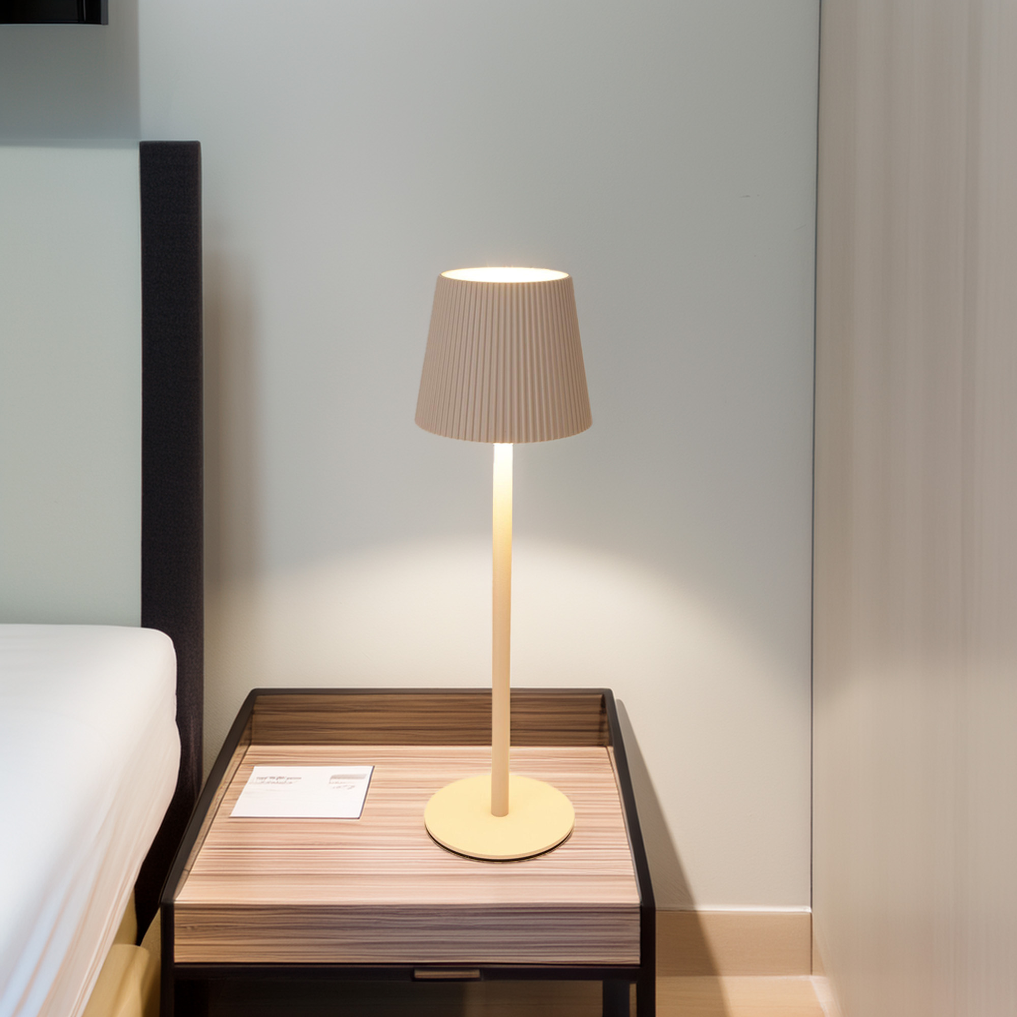 Una lámpara encendida en una mesita de noche. La lámpara tiene una pantalla y un soporte beiges. Una cama y una pared al fondo.