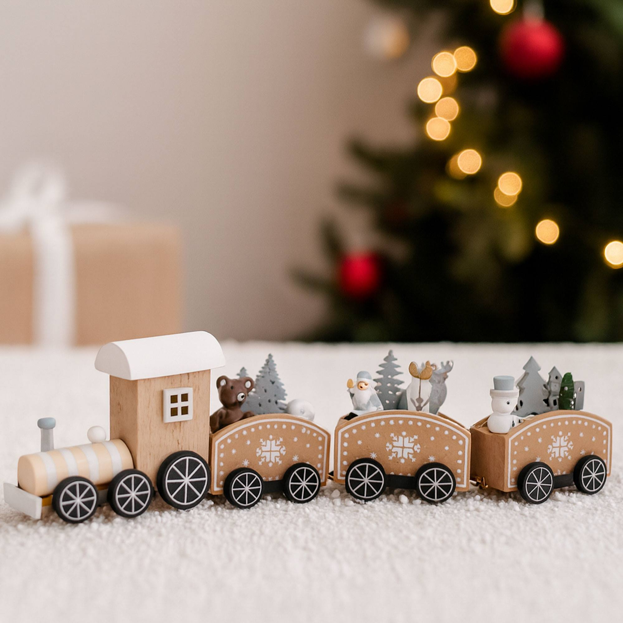 Un tren navideño de madera con figuras de oso, Papá Noel y muñeco de nieve, sobre una superficie blanca, con un árbol borroso de fondo.