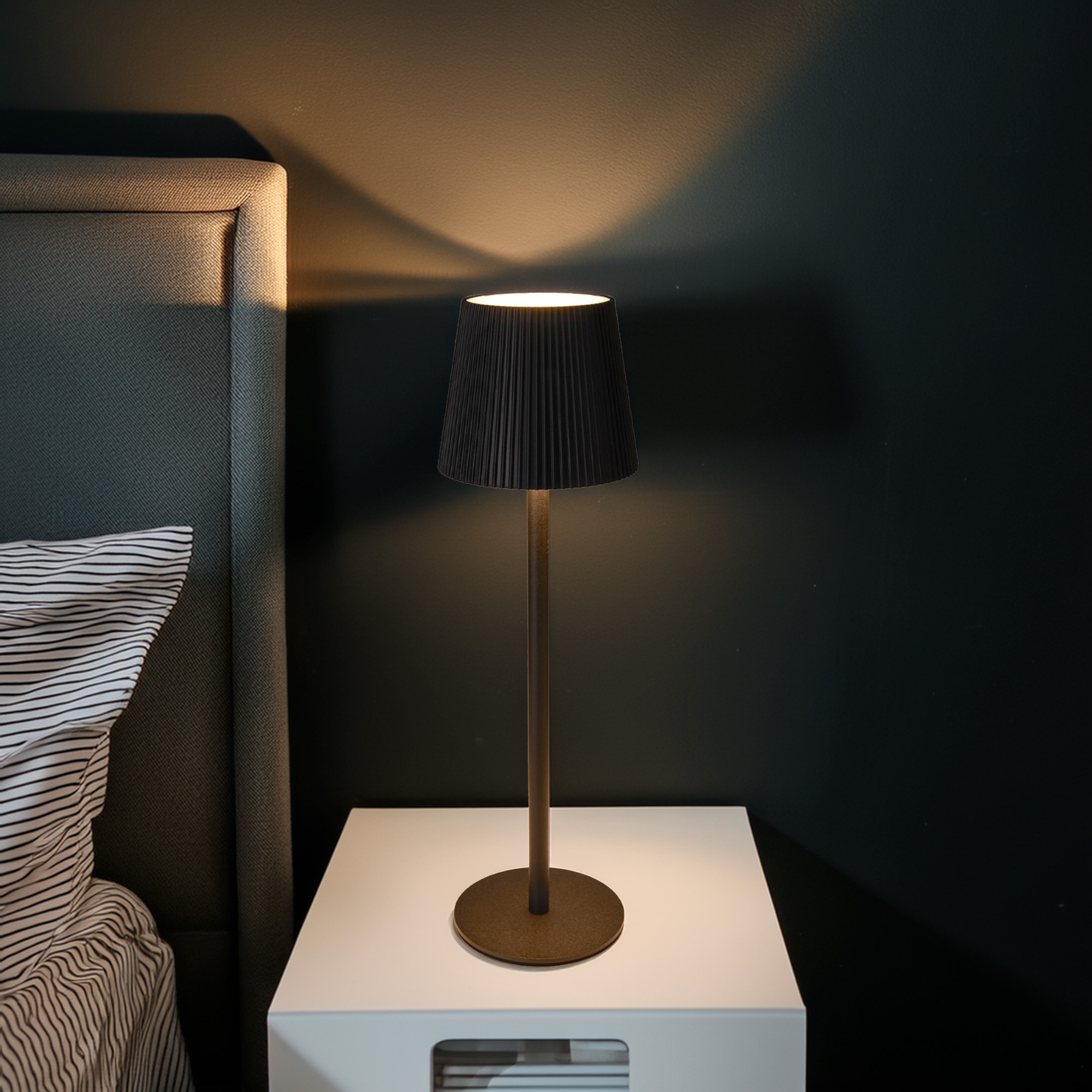 Una escena de mesita de noche con una lámpara sobre una mesa blanca. Una pantalla negra emite luz cálida. Cabecero gris y pared oscura.