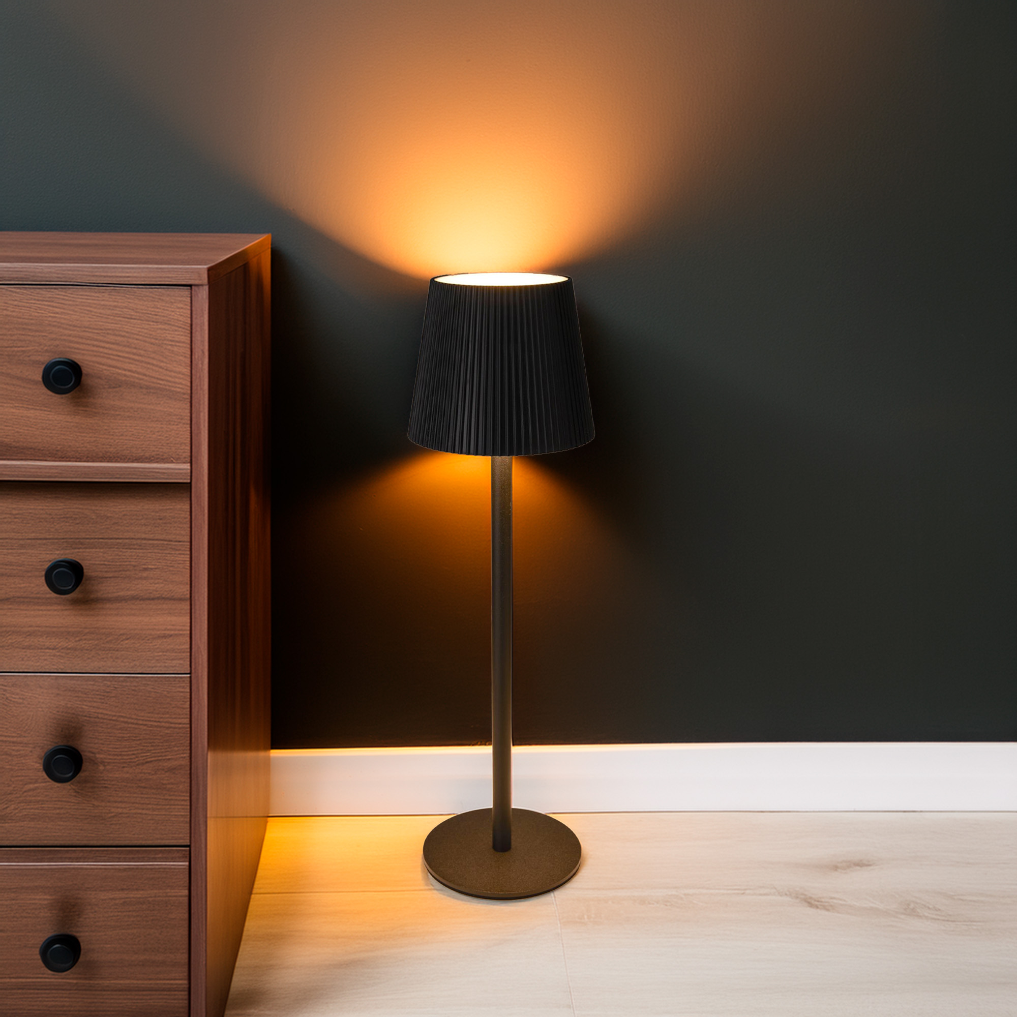 Una lámpara con pantalla negra ilumina una pared oscura. Un aparador de madera a la izquierda. Suelo de madera clara.