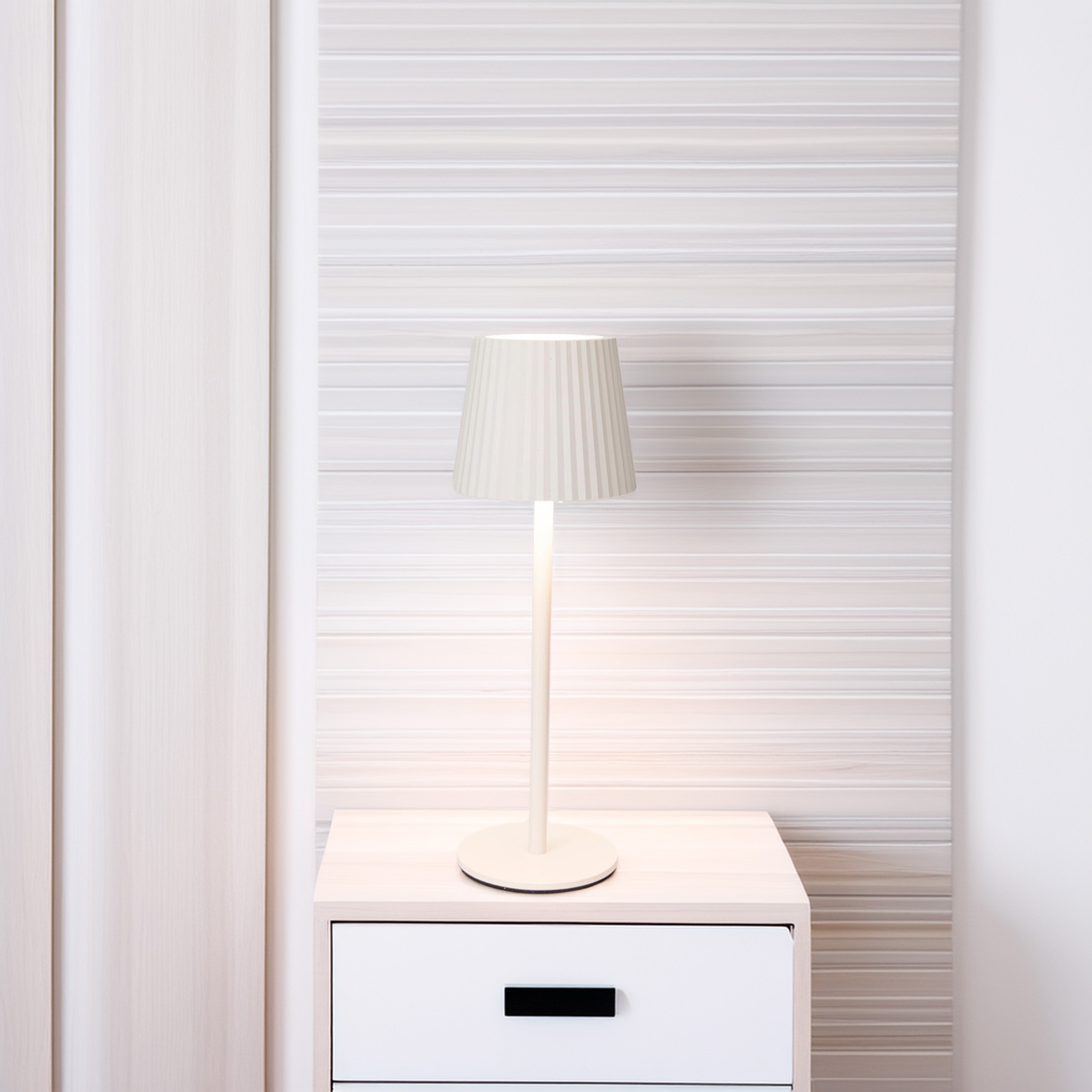 Una lámpara crema con pantalla en una mesita de noche blanca. Cortinas crema a la izquierda. Pared a rayas en el fondo.