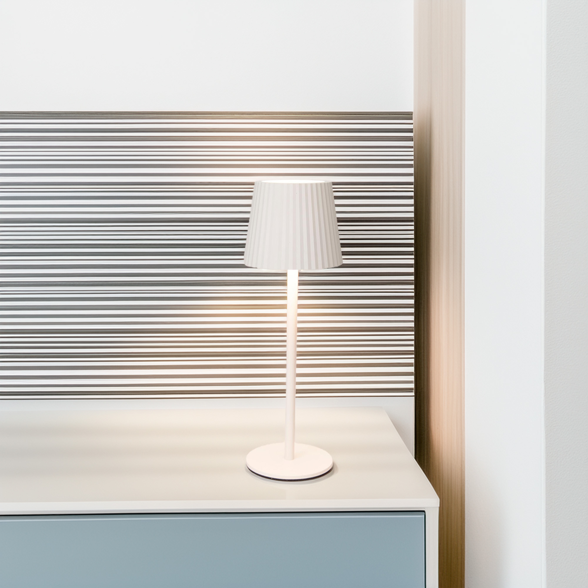 Una lámpara blanca con pantalla plisada sobre mesa. El fondo es rayado. A la derecha hay una pared blanca.