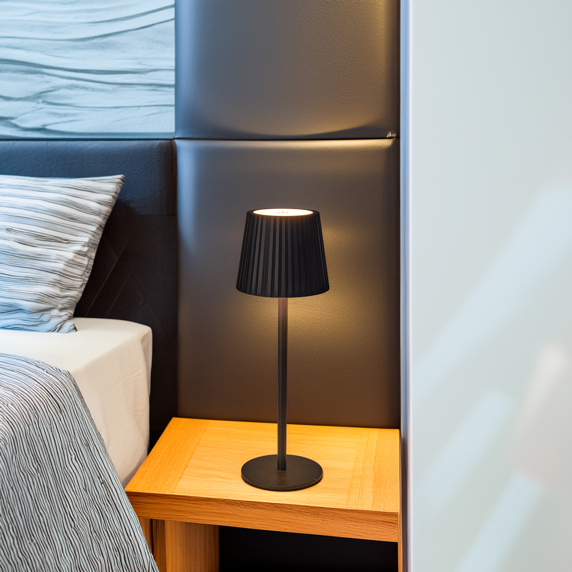 Una lámpara negra sobre una mesita de noche de madera junto a una cama con ropa de cama a rayas y un cabecero gris.