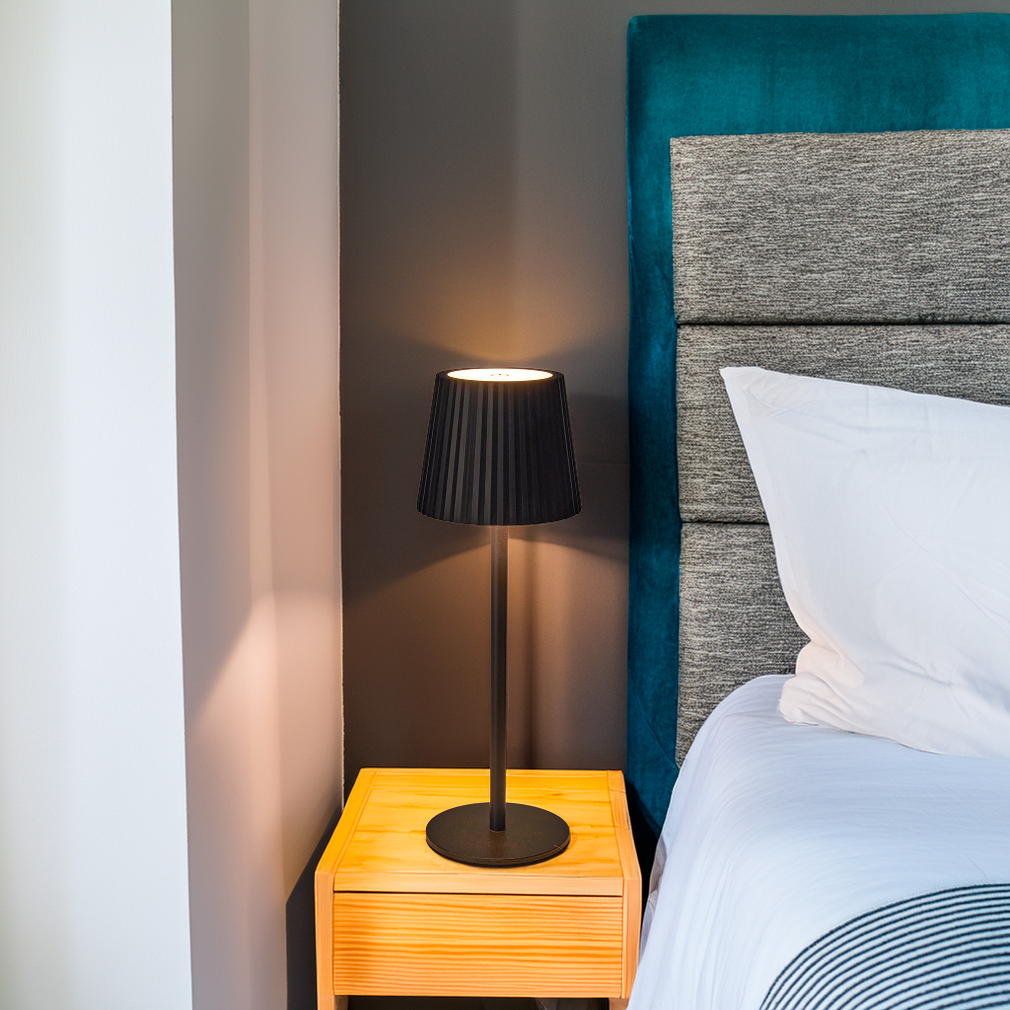 Una lámpara negra encendida sobre una mesita de noche de madera, junto a una cama con almohada blanca y cabecero turquesa.