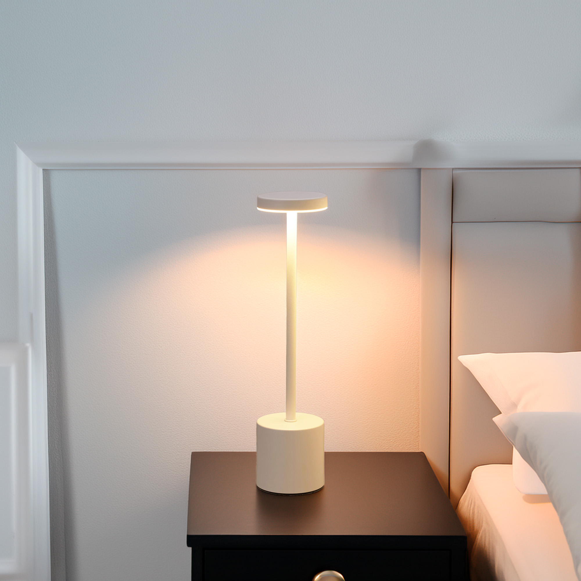 Una lámpara de noche blanca sobre una mesita de noche negra junto a una cama con almohadas blancas.