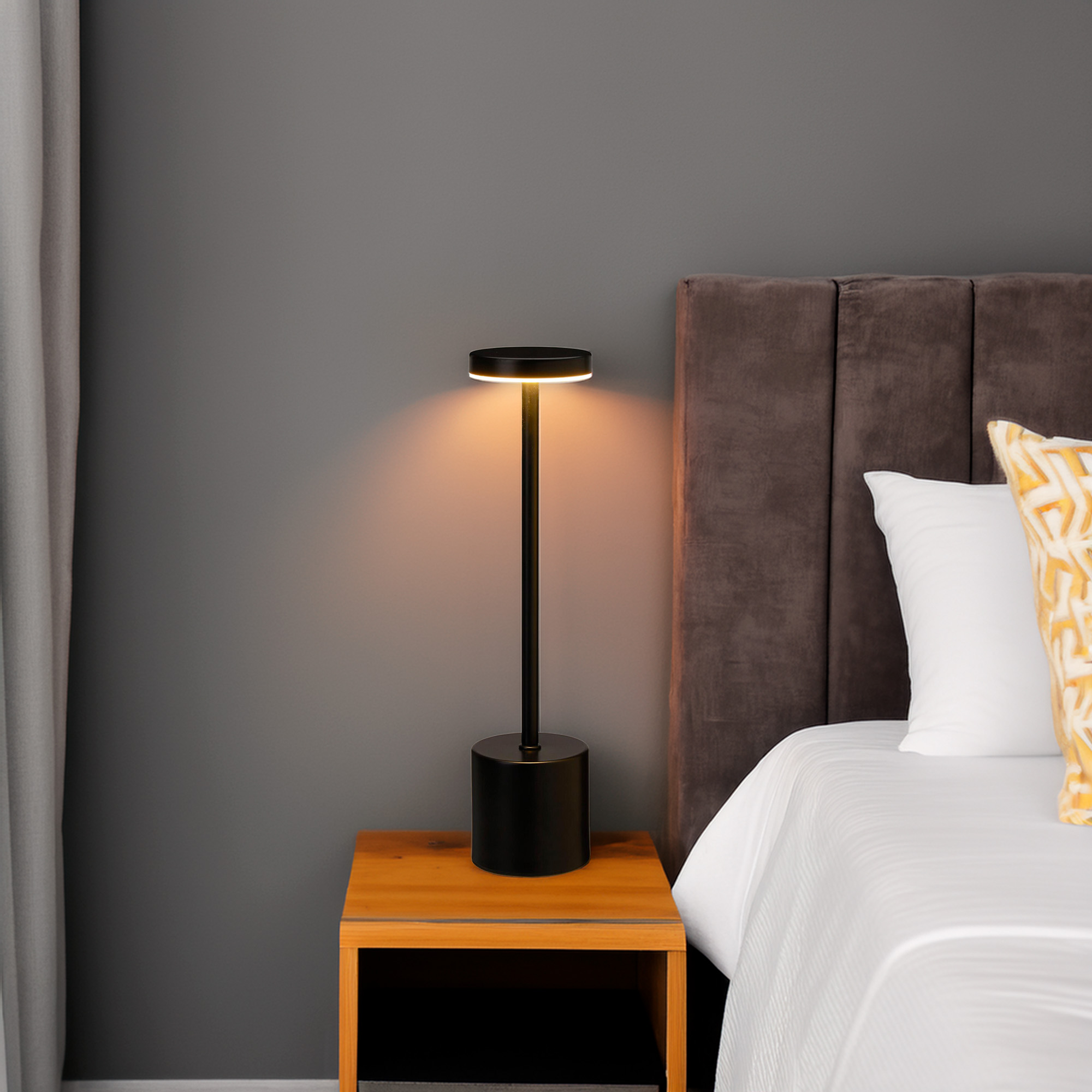 Una lámpara negra sobre una mesa de madera junto a una cama con sábanas blancas y una almohada estampada. Pared gris al fondo.