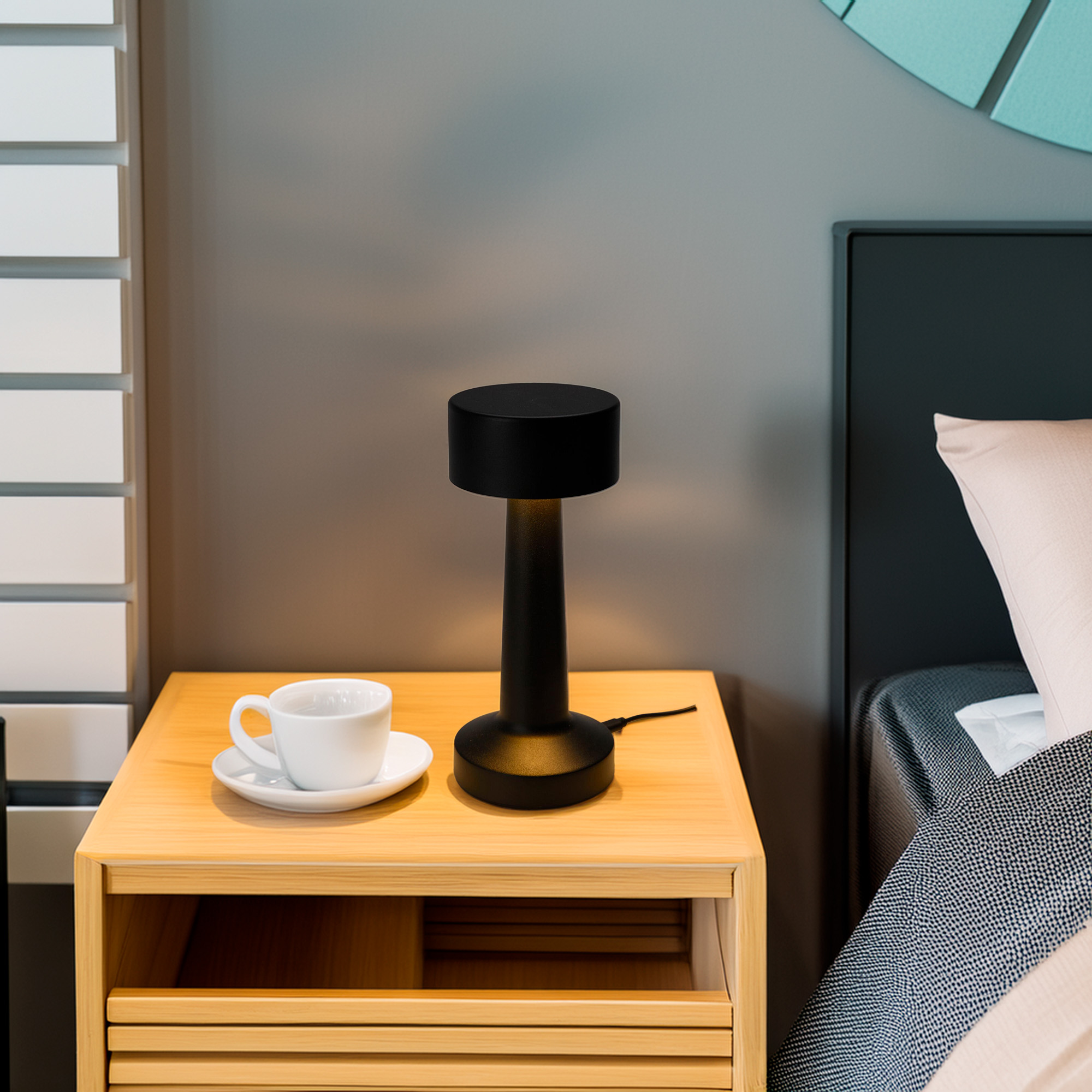 Una mesita de noche con lámpara y taza de café. Una cama y una persiana están al fondo.