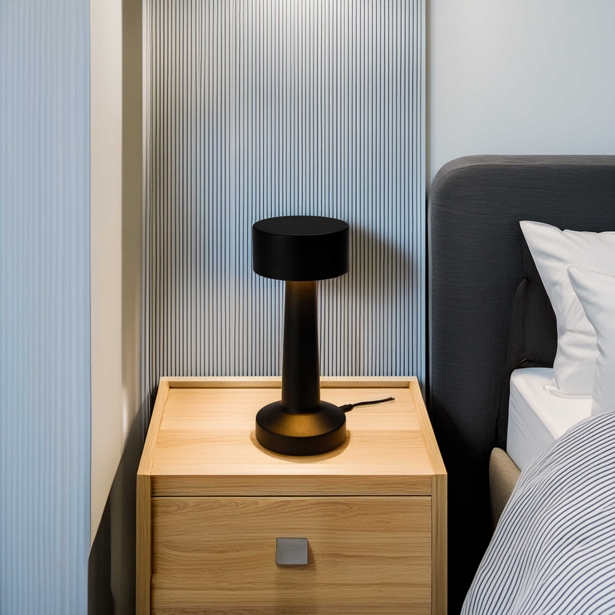Una lámpara negra sobre una mesita de noche de madera. Una cama con almohadas blancas a la derecha. Pared a rayas en el fondo.