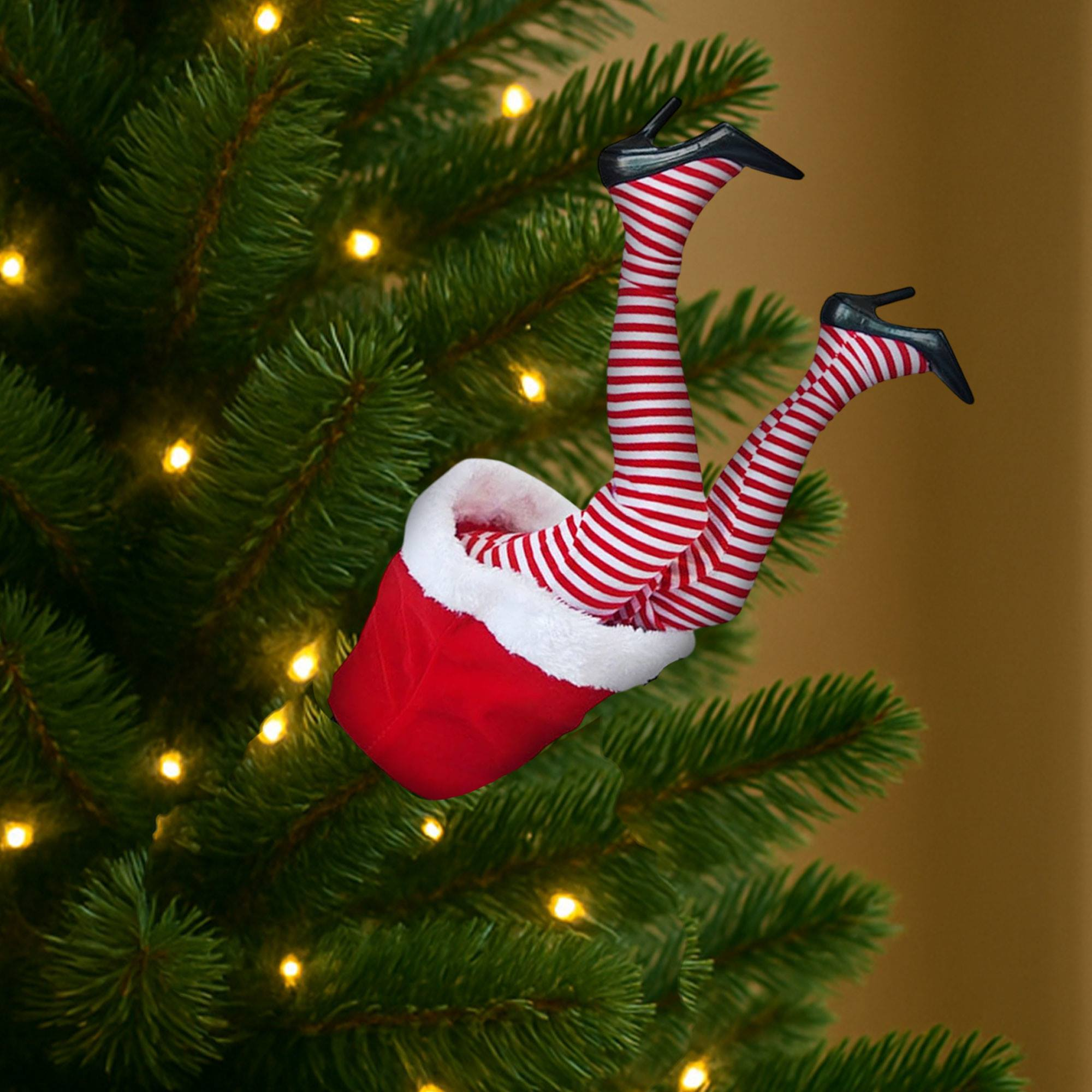 Piernas a rayas rojas y blancas con tacones negros emergen de un calcetín navideño en un árbol, con luces.