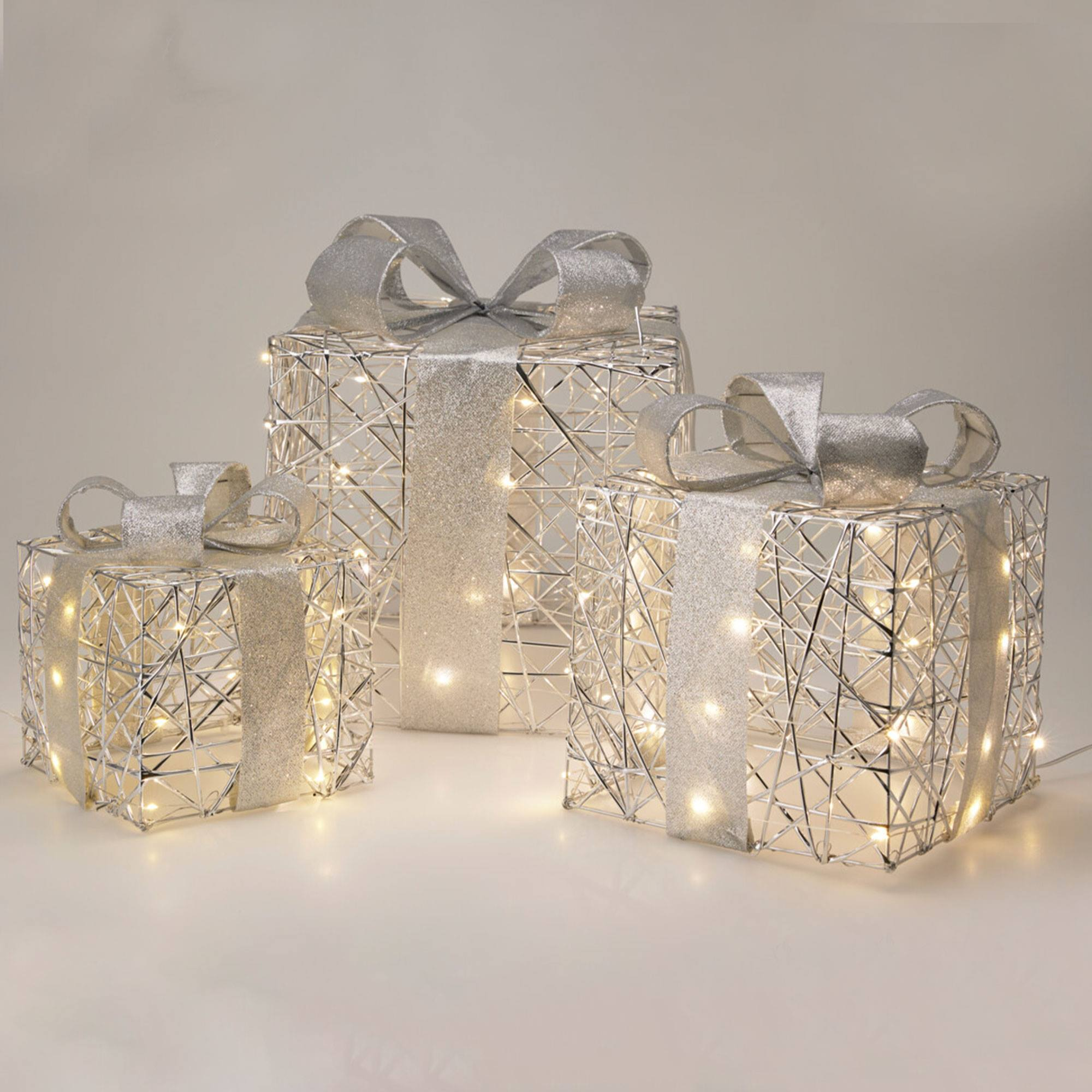 Tres cajas de regalo plateadas con lazos y cintas, iluminadas con luces de hadas, sobre una superficie blanca.