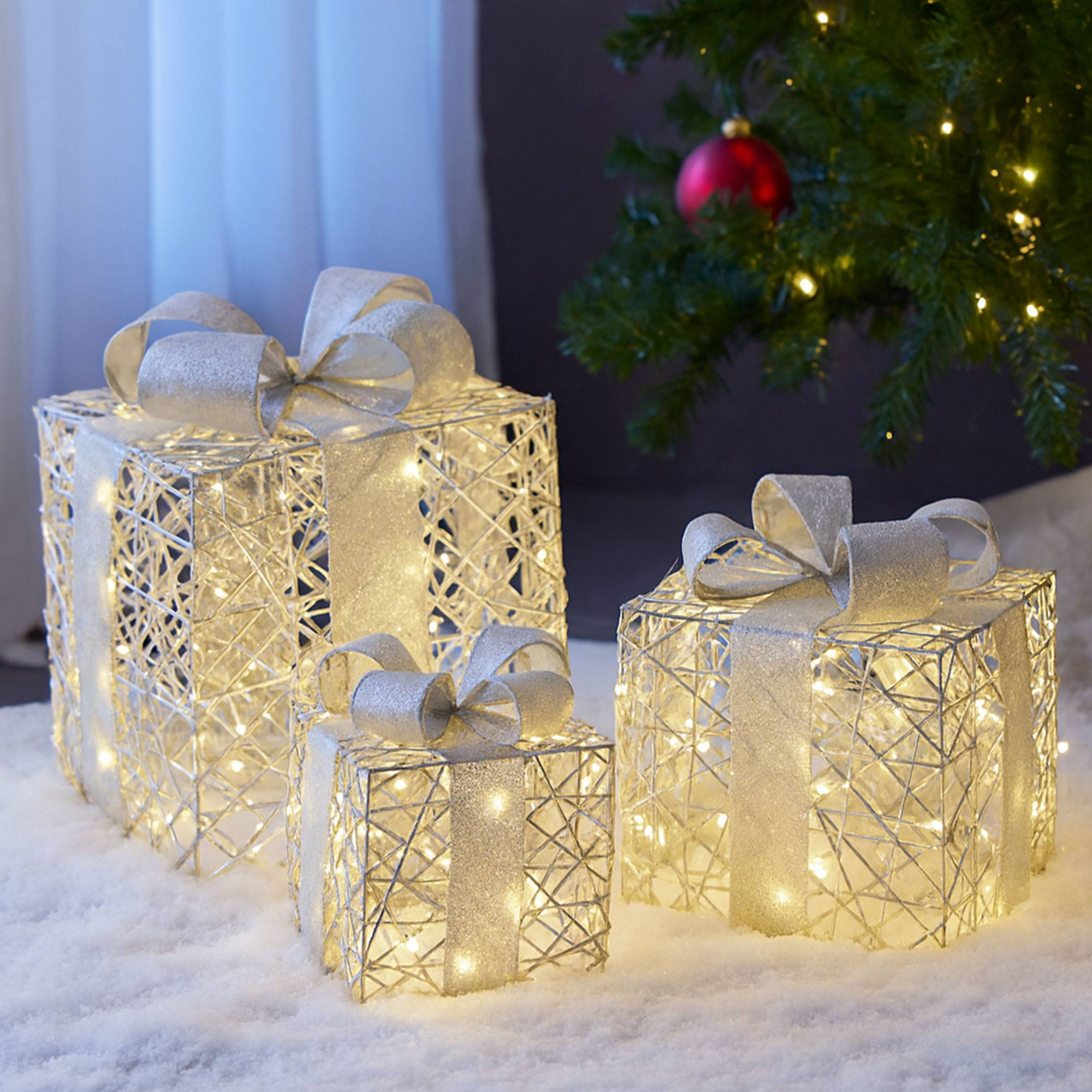 Tres cajas de regalo iluminadas con lazos plateados sobre una superficie nevada. Un árbol de Navidad y un adorno rojo al fondo.