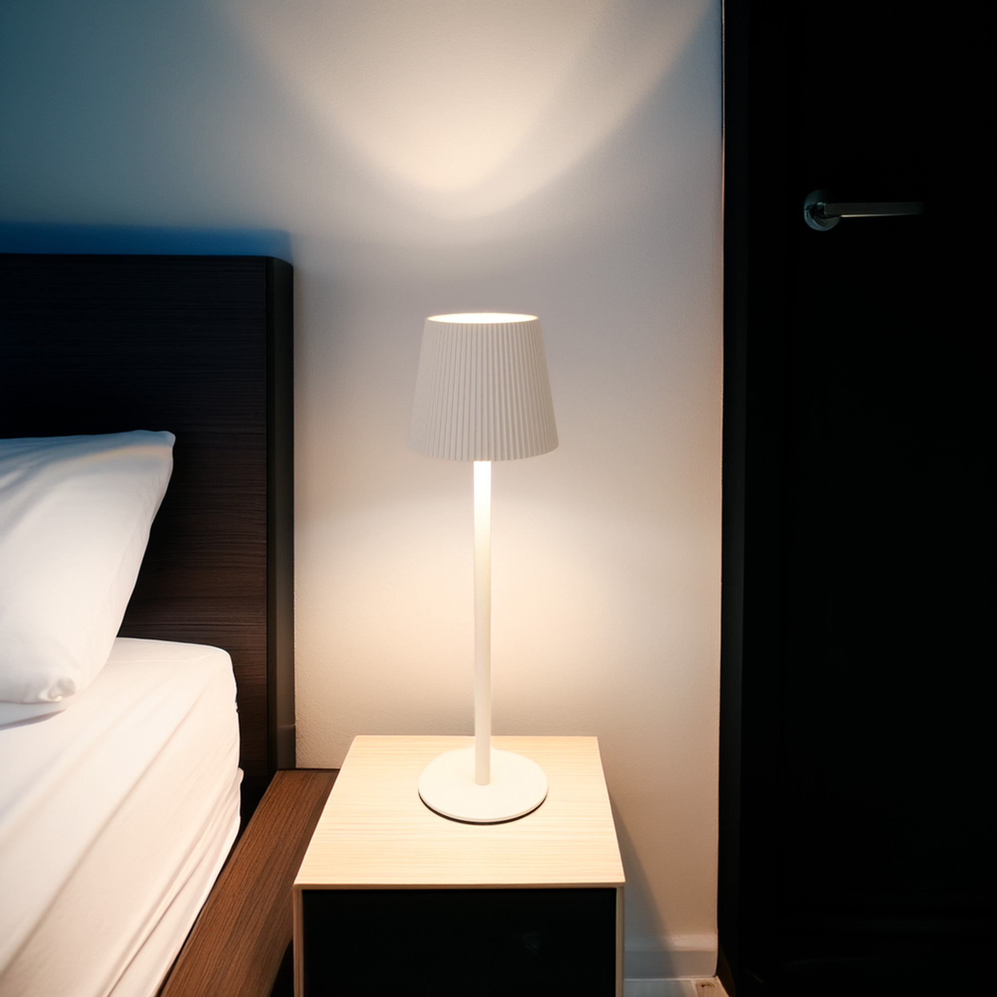 Una lámpara blanca sobre una mesita de noche de madera. Marco de cama a la izquierda. Puerta negra a la derecha.