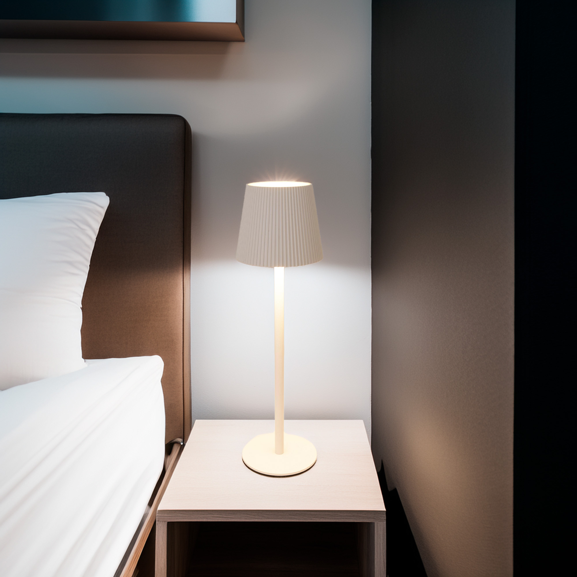 Una lámpara encendida está sobre una mesita de noche junto a una cama. La lámpara tiene una pantalla crema. La cama tiene un edredón blanco.