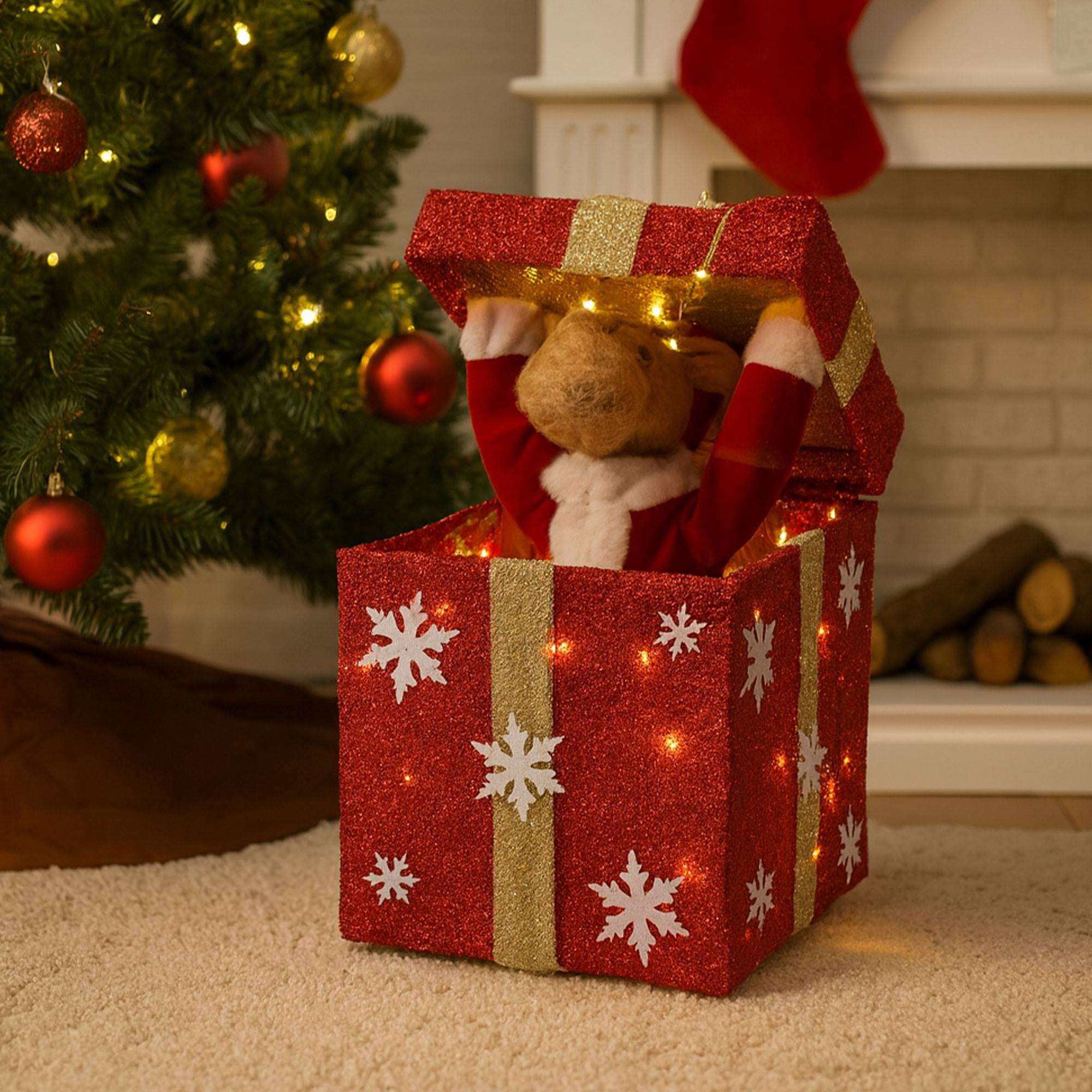 Un reno en una caja de regalo roja con purpurina, copos de nieve y luces. Se ve un árbol de Navidad con adornos.