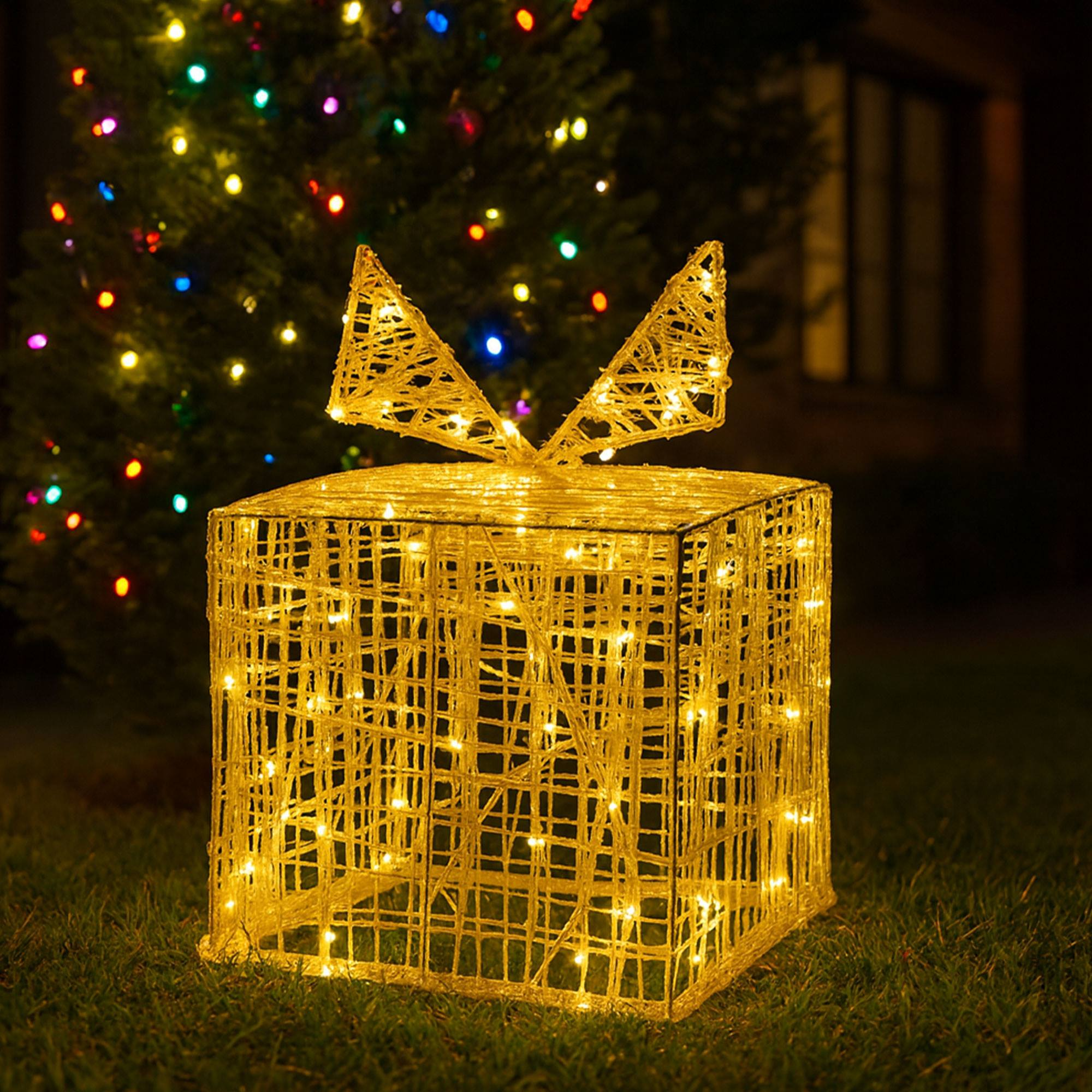 Una caja de regalo navideña iluminada con lazo, en un jardín. Luces en un árbol al fondo. Es de noche.
