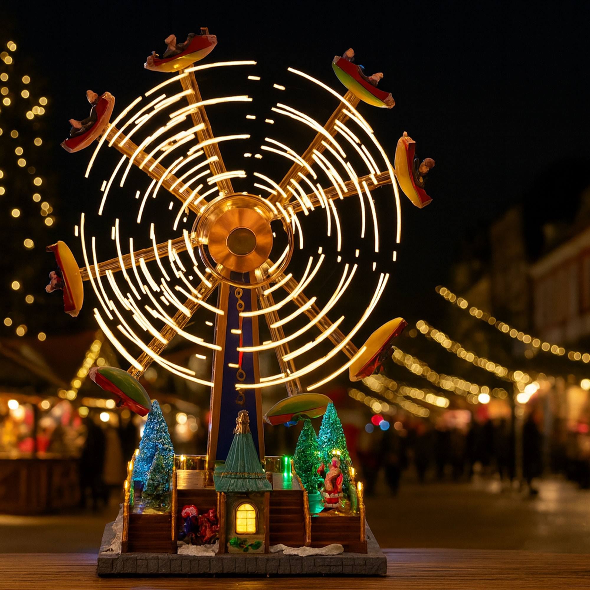 Una noria en miniatura iluminada con pequeñas figuras, árboles y Papá Noel. El fondo tiene luces borrosas.