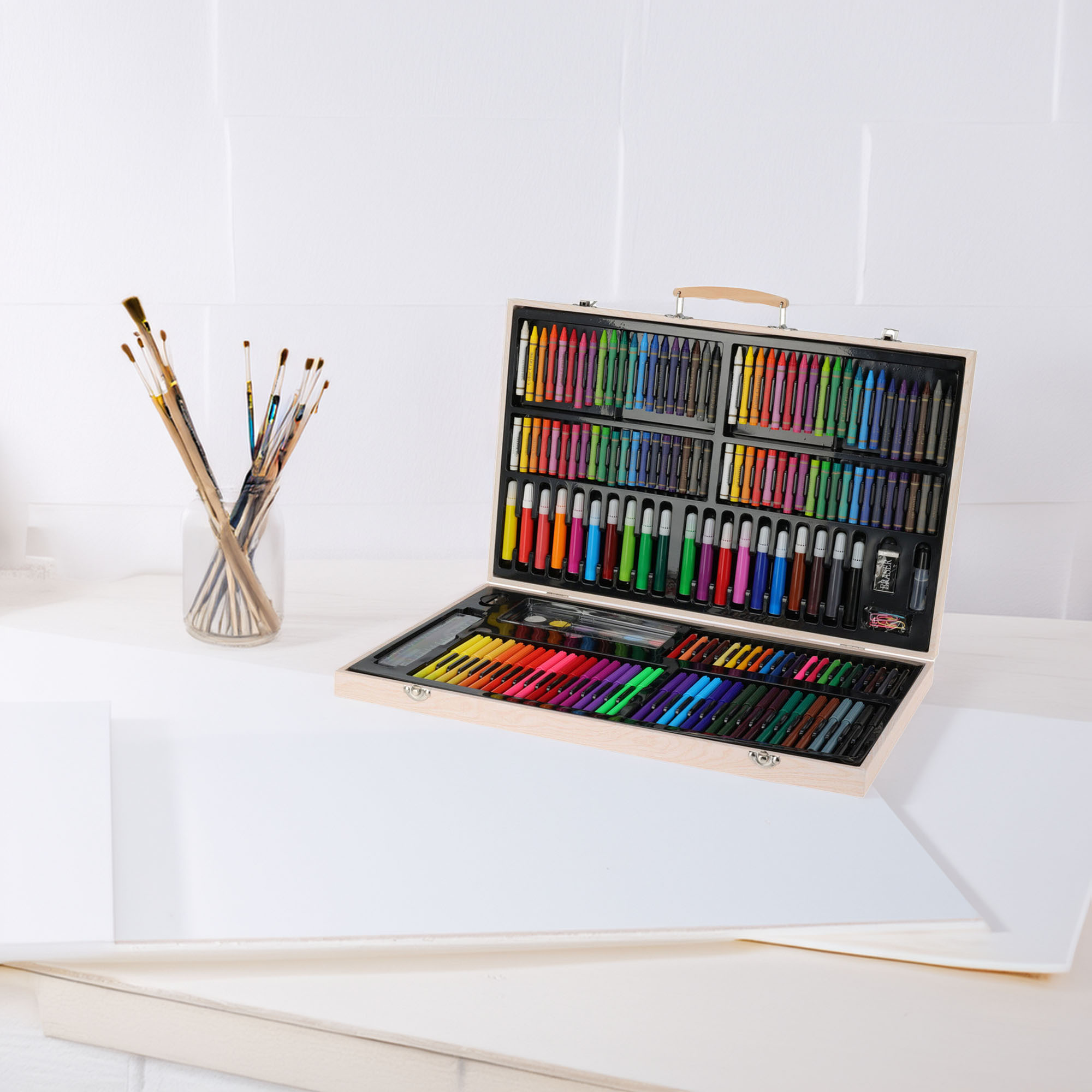 Kit de arte abierto con crayones, marcadores y lápices. Pinceles en un frasco a la izquierda. Hojas de papel blanco sobre la mesa.