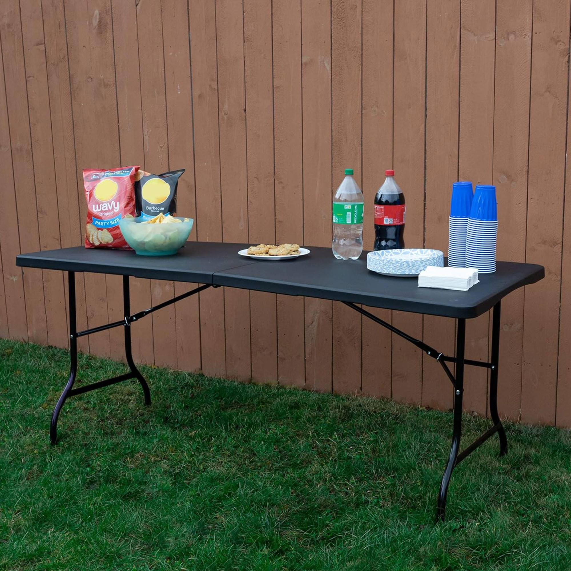 Una mesa con patatas fritas, galletas, bebidas y vasos se encuentra sobre la hierba frente a una valla de madera marrón.