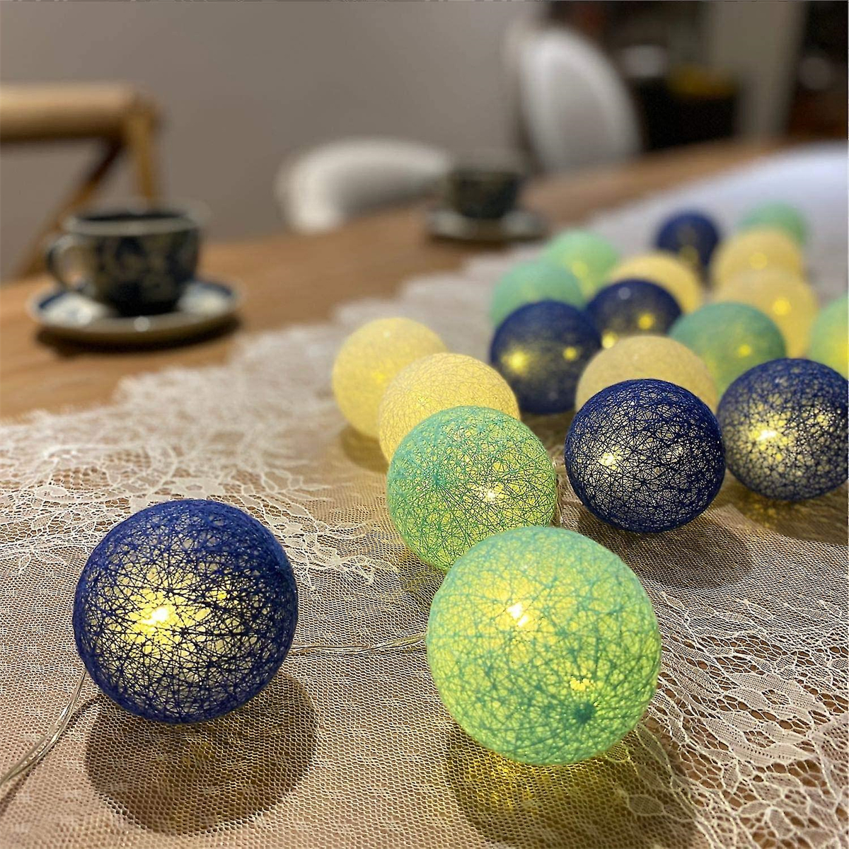 Lichterketten mit gelben, grünen und blauen Kugeln auf einem Tisch mit weißem Spitzentischläufer. Im Hintergrund eine Teetasse.