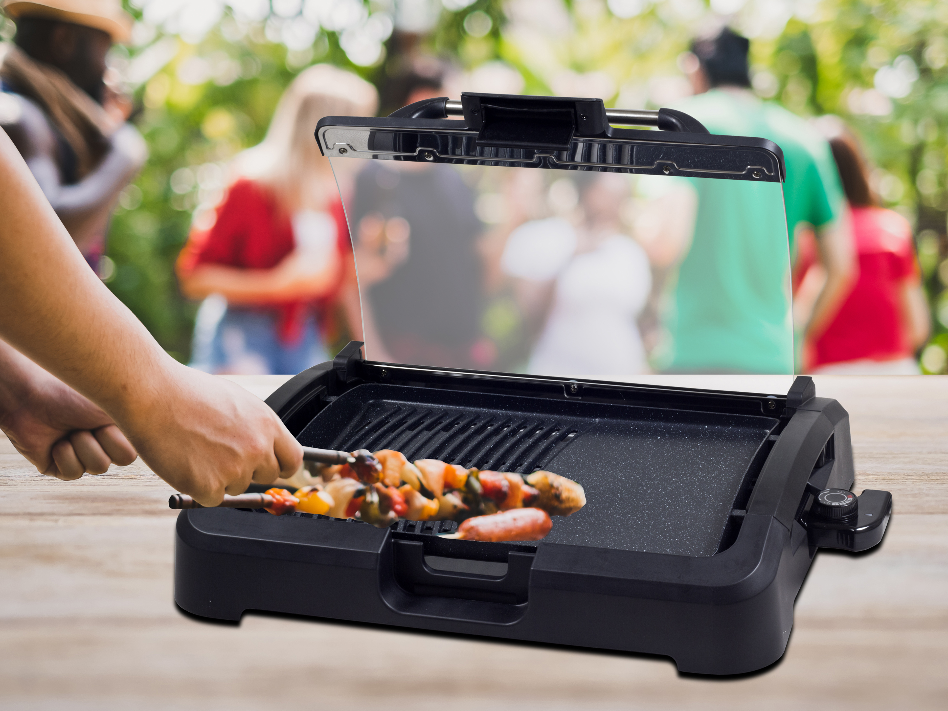 Hände grillen Spieße auf einem schwarzen Grill. Personen sind im Hintergrund verschwommen, ein Grillfest findet statt.