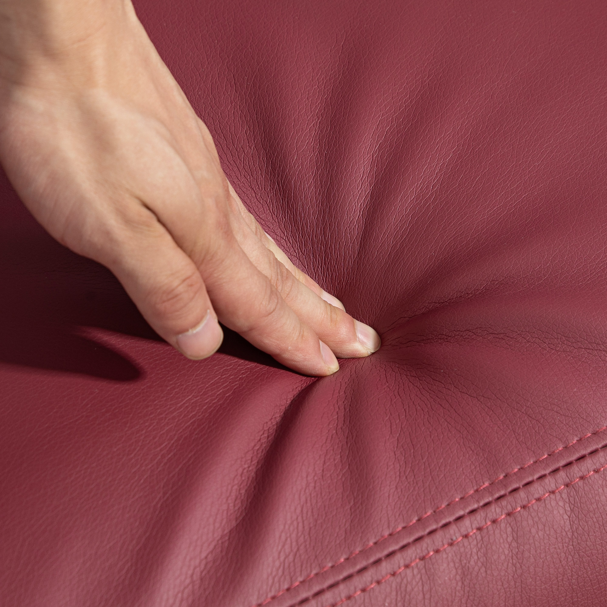 La mano preme sulla pelle bordeaux, creando un'ammaccatura. Cuciture rosse visibili.