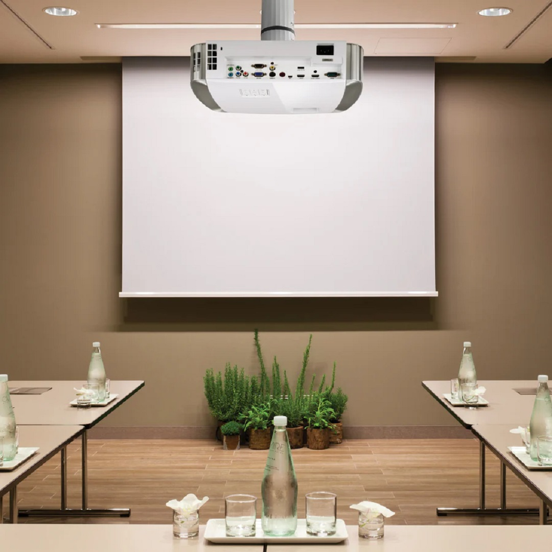 Sala de conferencias con proyector, pantalla, mesas, botellas de agua y plantas. Paredes beiges y suelo de madera.