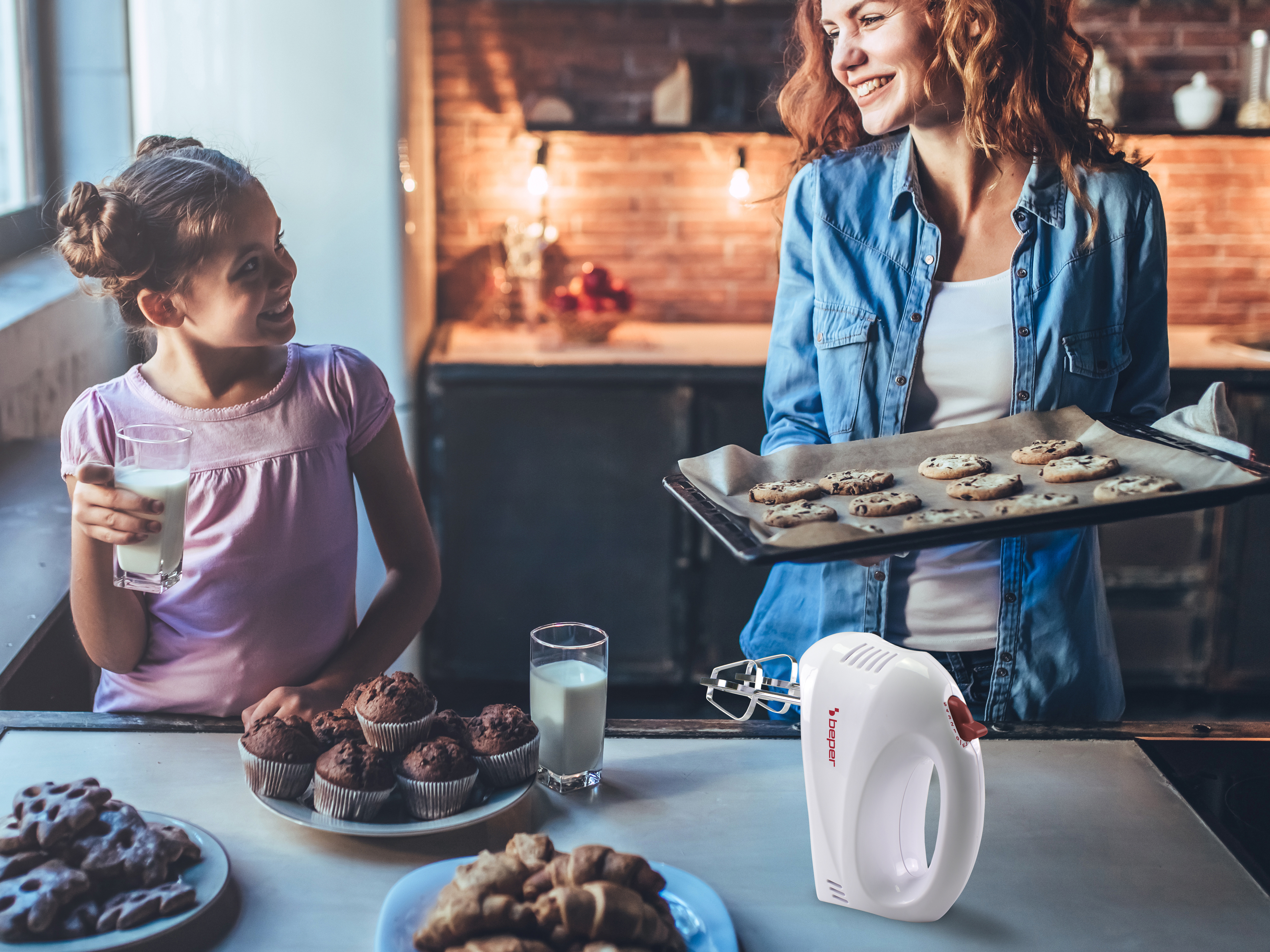Una donna con biscotti, una ragazza con latte e prodotti da forno sul tavolo. Un frullatore sul bancone.