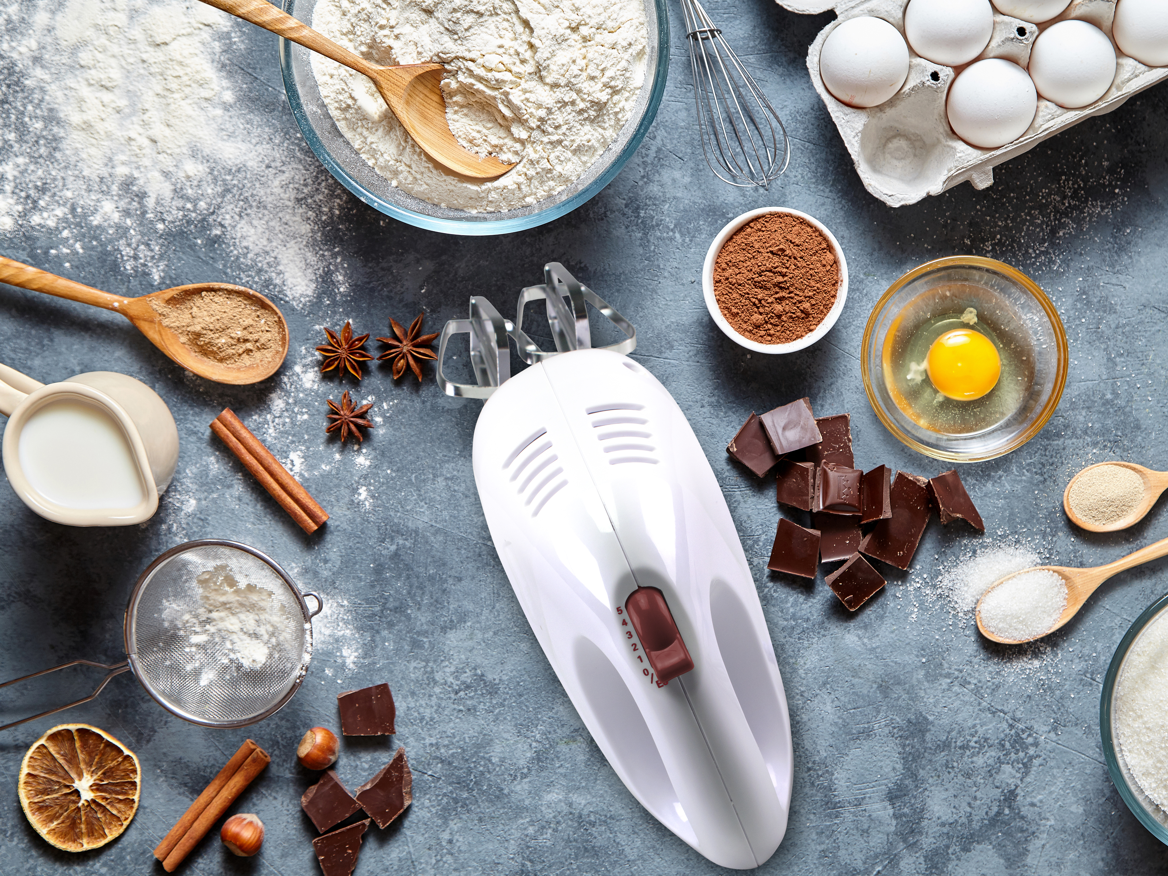 Vista dall'alto degli ingredienti per la cottura: farina, uova, cioccolato, cacao, spezie e sbattitore su superficie grigia.