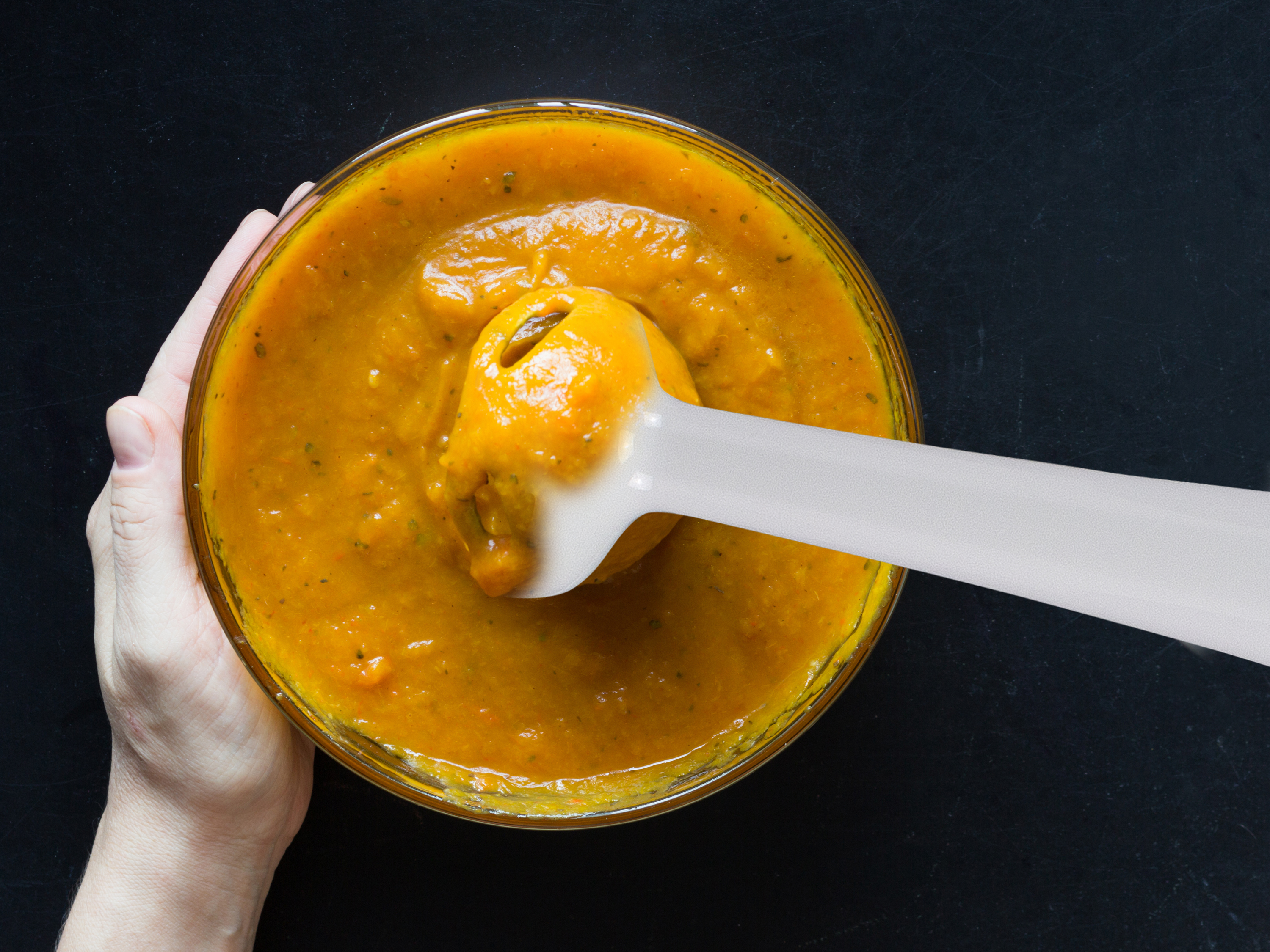 Widok z góry na dłoń trzymającą szklaną miskę z pomarańczową zupą i biały blender ręczny.