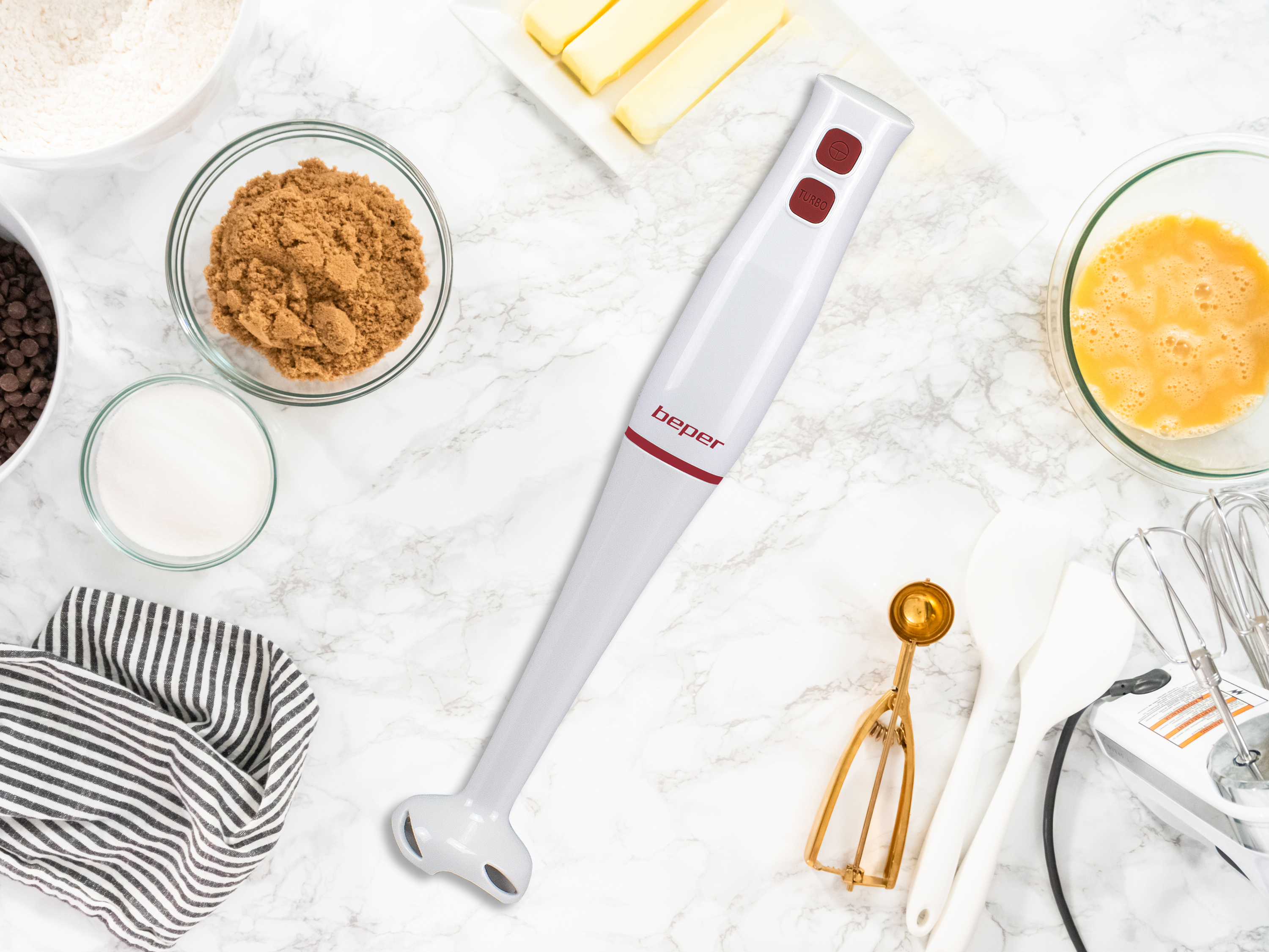 Widok z góry na składniki i narzędzia do pieczenia: mąka, cukier, brązowy cukier, kawałki czekolady, jajka, masło i blender.