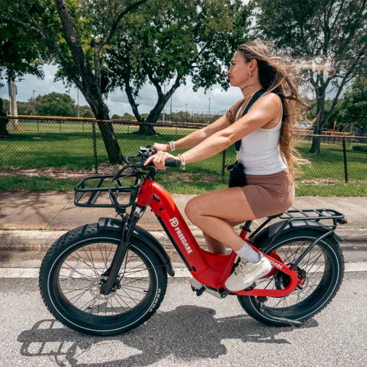 Kobieta jedzie na czerwonym rowerze elektrycznym po drodze. Ma na sobie biały top i szorty. W tle drzewa i park.