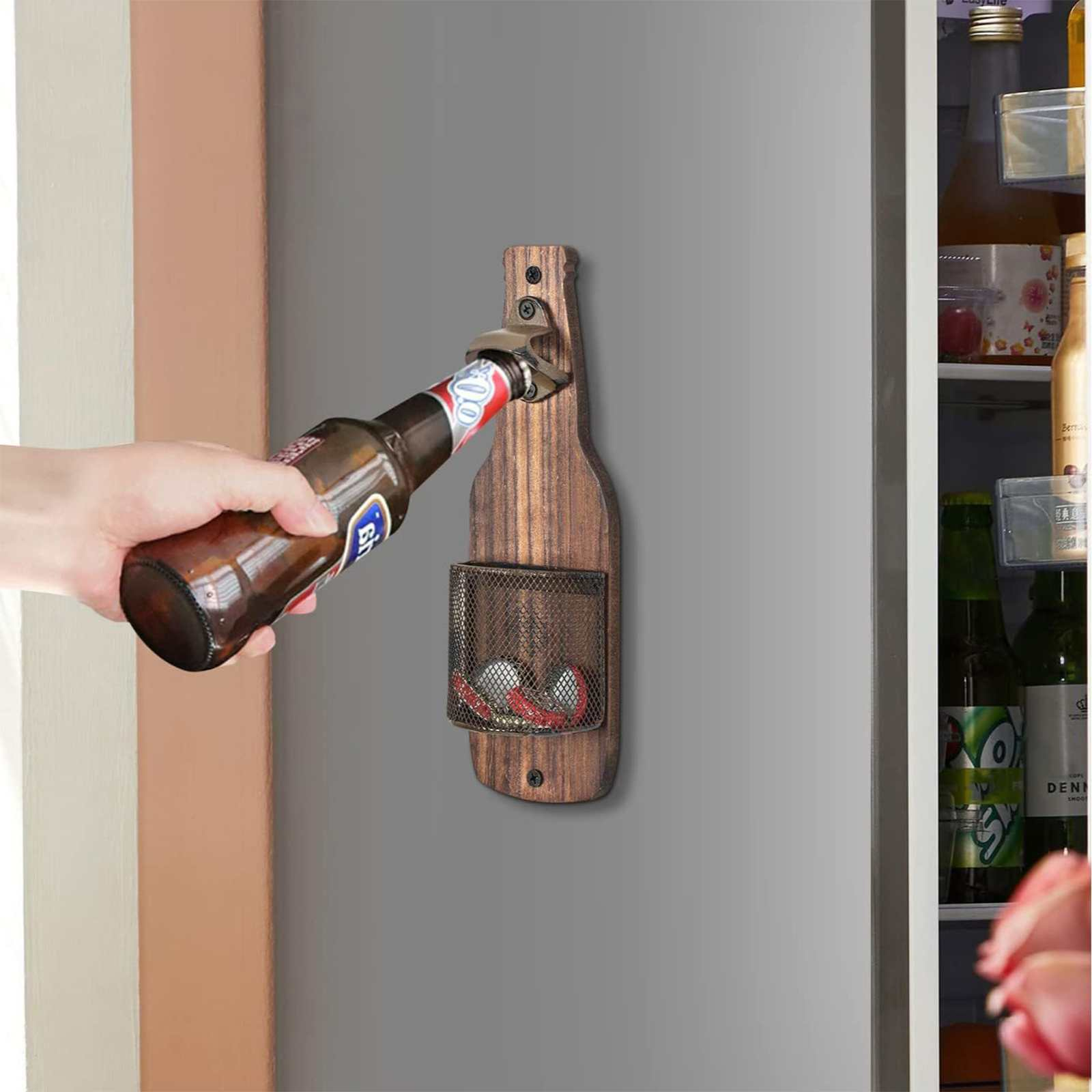 Eine Hand öffnet eine Bierflasche mit einem Flaschenöffner, der an einem Holzhalter befestigt ist. Ein Kühlschrank ist im Hintergrund.