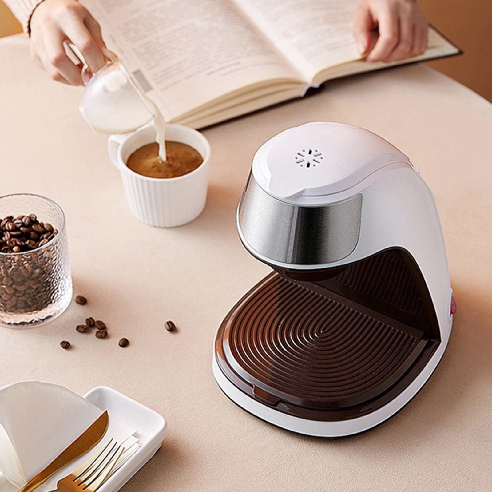 Eine Person gießt Milch in Kaffee, neben einer Kaffeemaschine, Kaffeebohnen und einem Buch.
