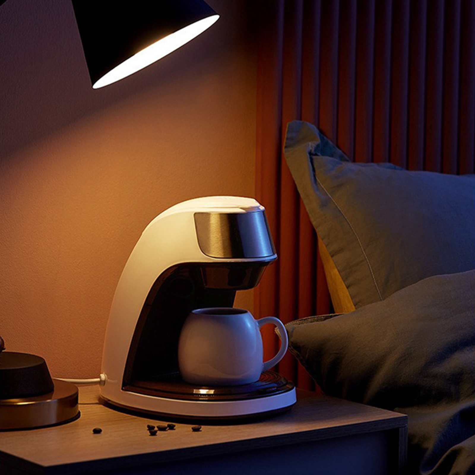 Eine Kaffeemaschine mit einer Tasse steht auf einem Nachttisch, beleuchtet von einer Lampe. Im Hintergrund ein Bett mit Kissen.