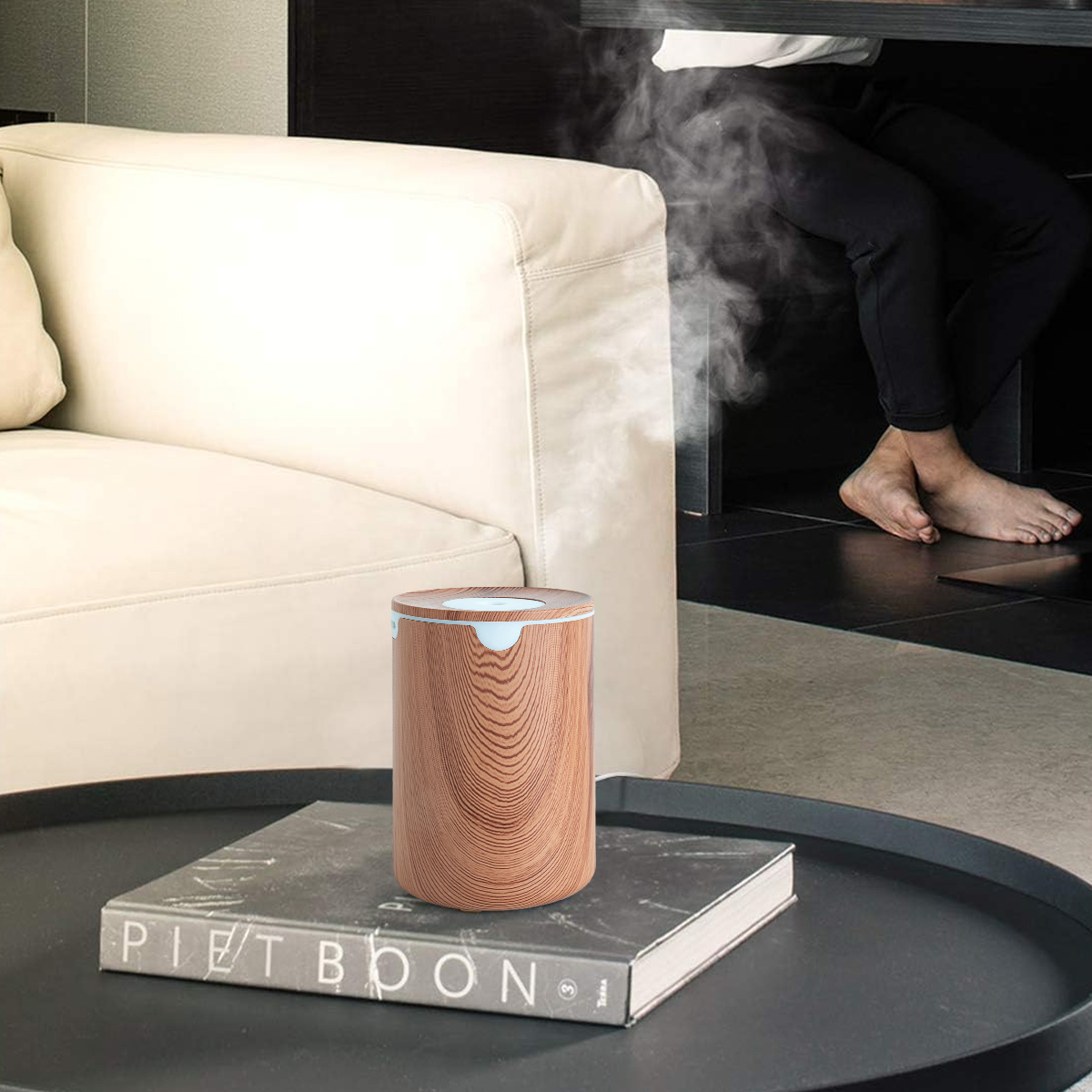 Ein Holzluftbefeuchter auf einem Tisch mit einem Buch. In der Nähe sitzt eine Person. Weißes Sofa im Hintergrund.