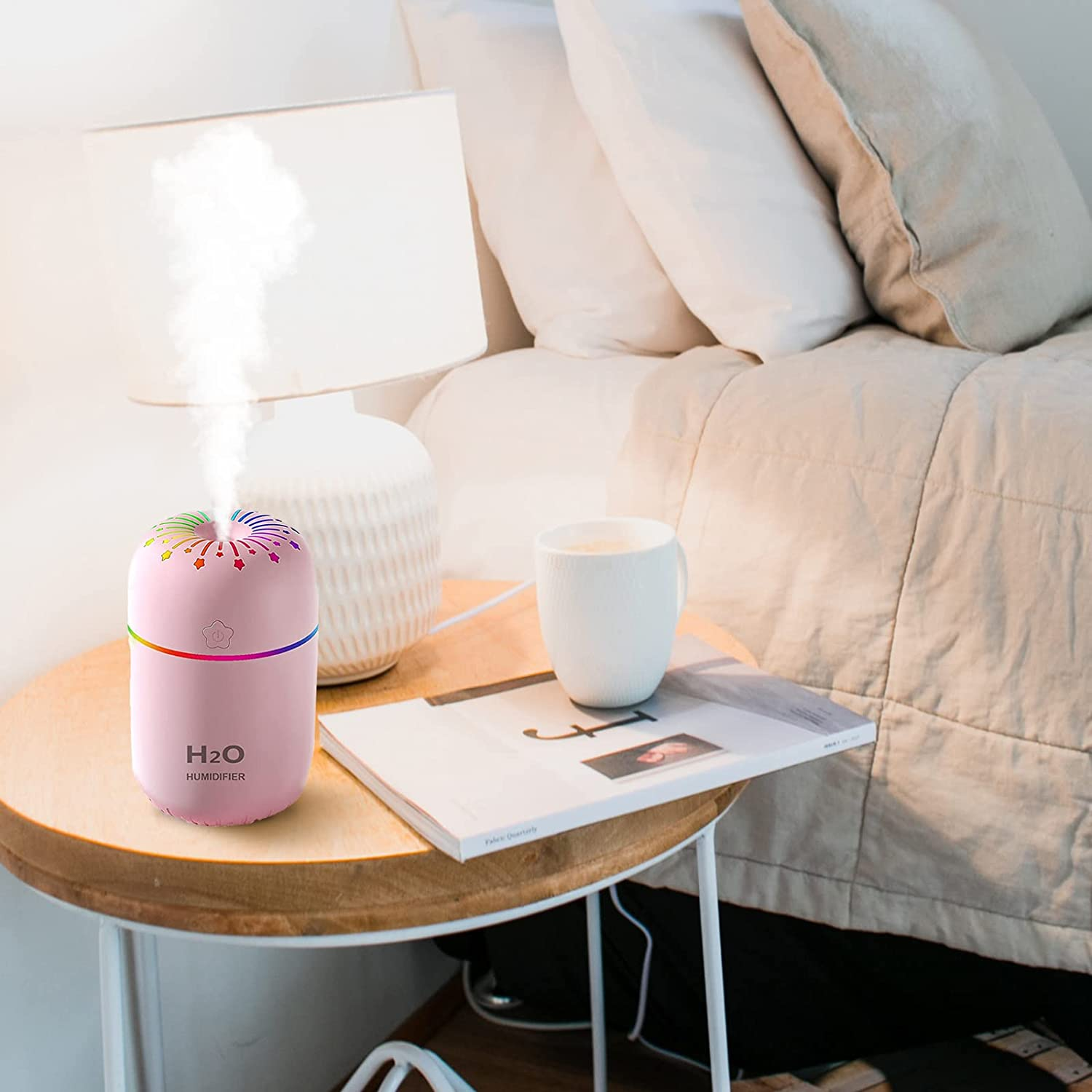 Ein rosa Luftbefeuchter mit Regenbogenlichtern gibt Dampf ab. Daneben stehen eine Lampe, eine weiße Tasse und eine Zeitschrift auf einem Holztisch.