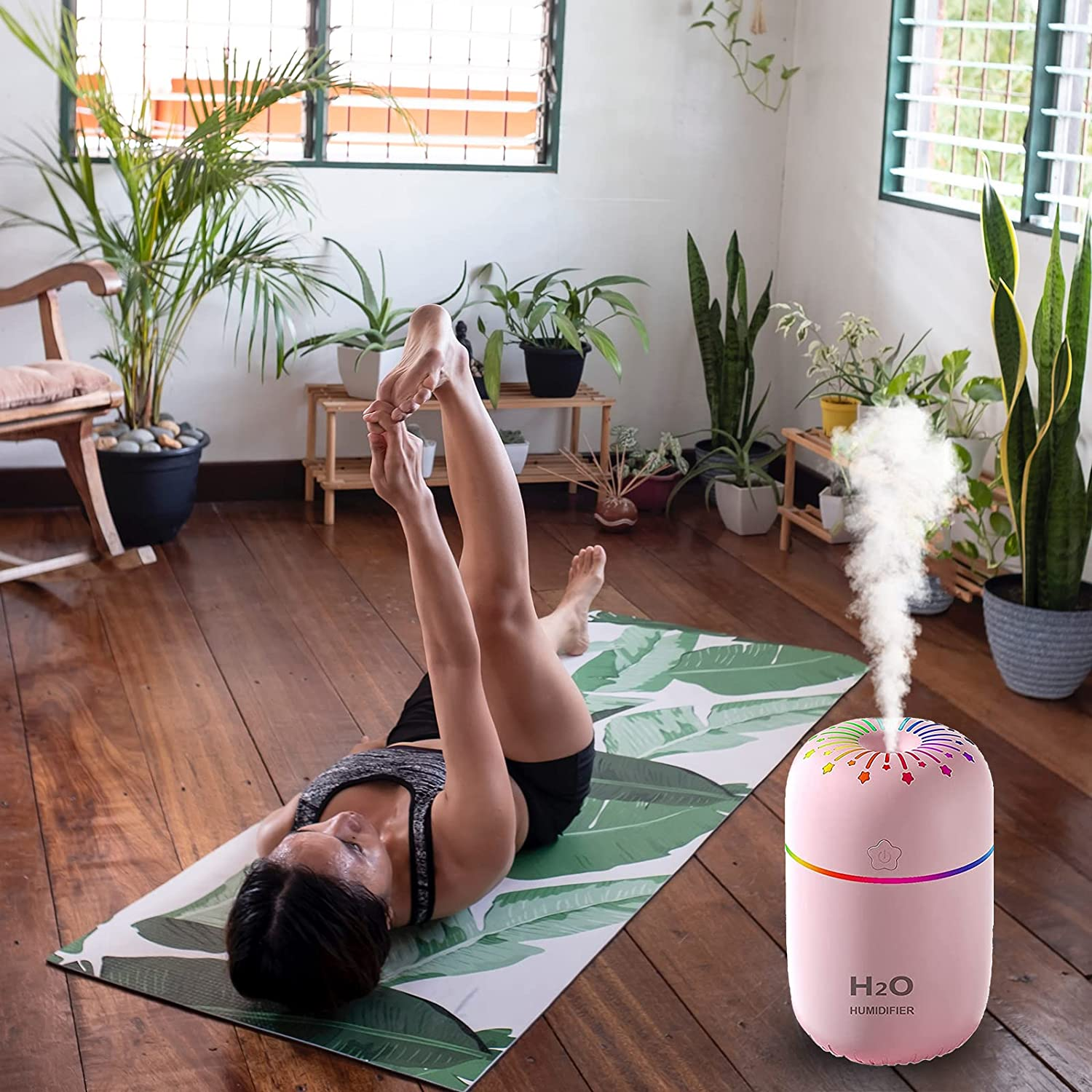 Frau macht Yoga auf einer Matte mit Blattmuster. Ein rosa Luftbefeuchter gibt Dampf ab. Pflanzen, Holzboden und Fenster sind im Hintergrund.