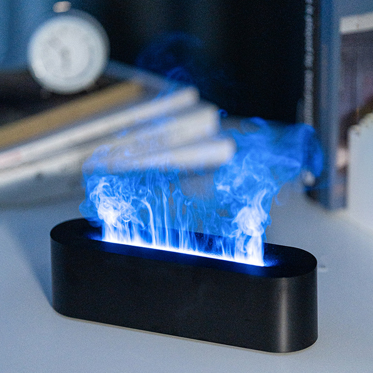 Ein schwarzes, ovalförmiges Gerät emittiert blau gefärbte simulierte Flammen. Im Hintergrund Bücher und eine weiße Uhr auf einem Tisch.