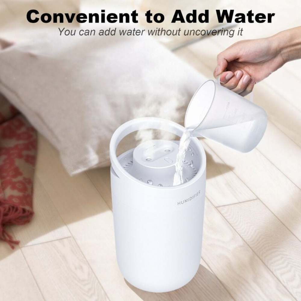 Eine Hand gießt Wasser in einen weißen Luftbefeuchter. Die Szene umfasst ein Kissen, einen Schal und Holzfußboden.