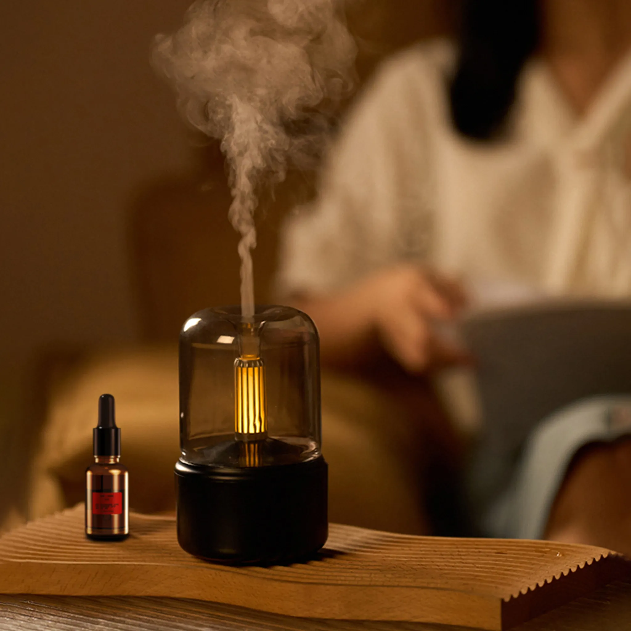 Ein Aromadiffusor gibt Dampf ab, neben einer Ölflasche. Im Hintergrund ist eine Person verschwommen, die liest.