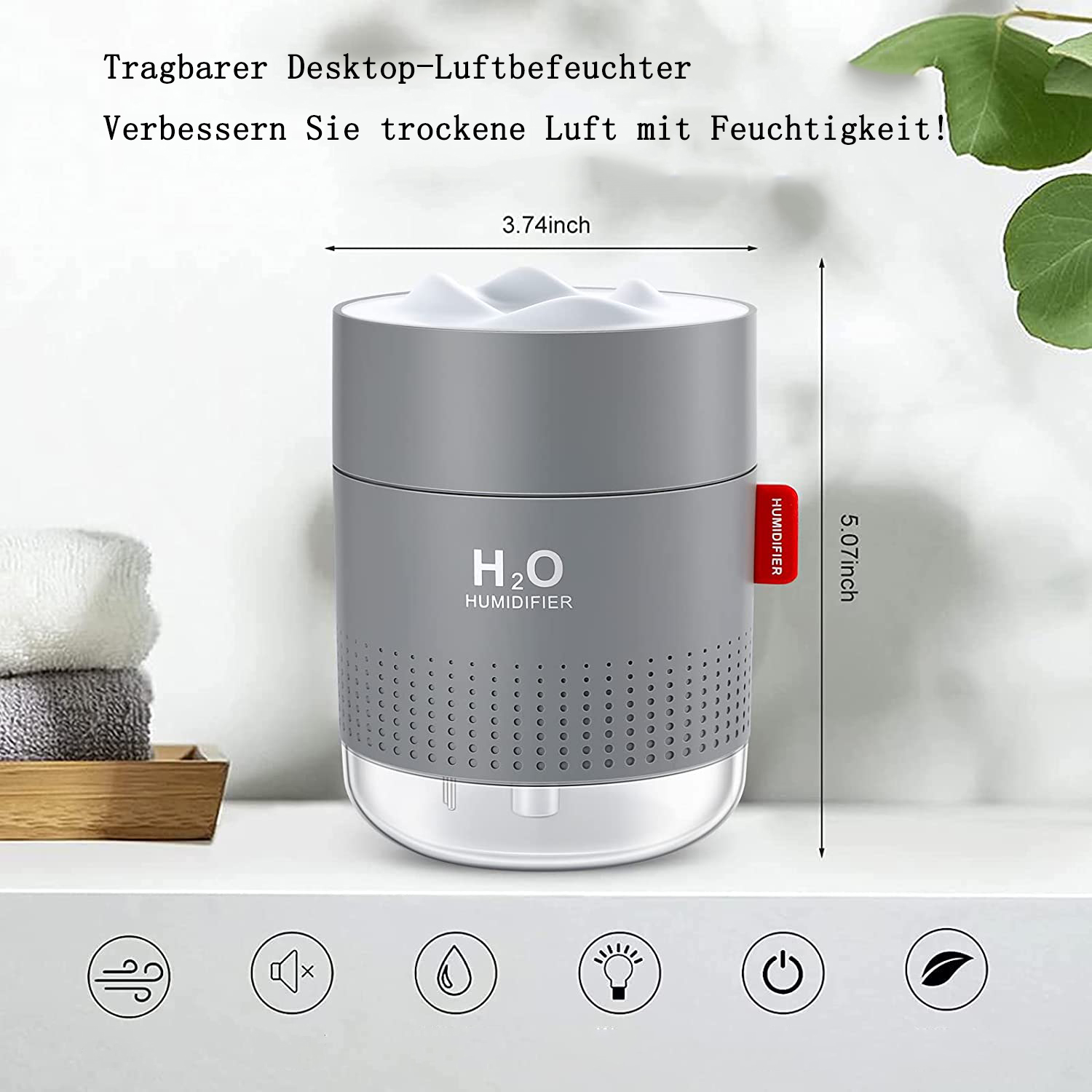 Ein grauer Luftbefeuchter mit weißer Oberseite. Er hat H2O darauf, ein rotes 'Luftbefeuchter'-Etikett und darunter Symbole. Ein Handtuch und ein Blatt sind im Hintergrund.