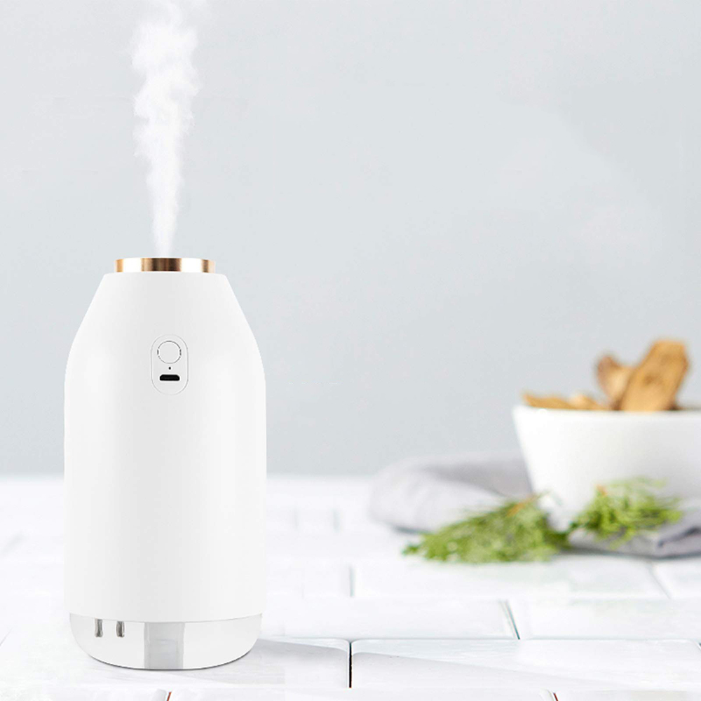 Weißer Aroma-Diffusor mit Dampf, Goldrand, auf weißen Fliesen. Schüssel mit Holzstücken im Hintergrund. Grün und ein Handtuch.