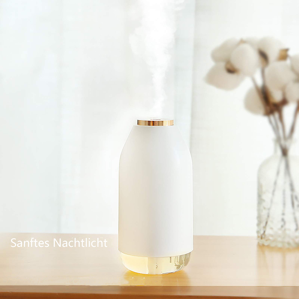 Ein weißer Aroma-Diffusor mit goldenem Rand verströmt Dampf. Er steht auf einem Holztisch, im Hintergrund weiße Blumen in einer Vase.