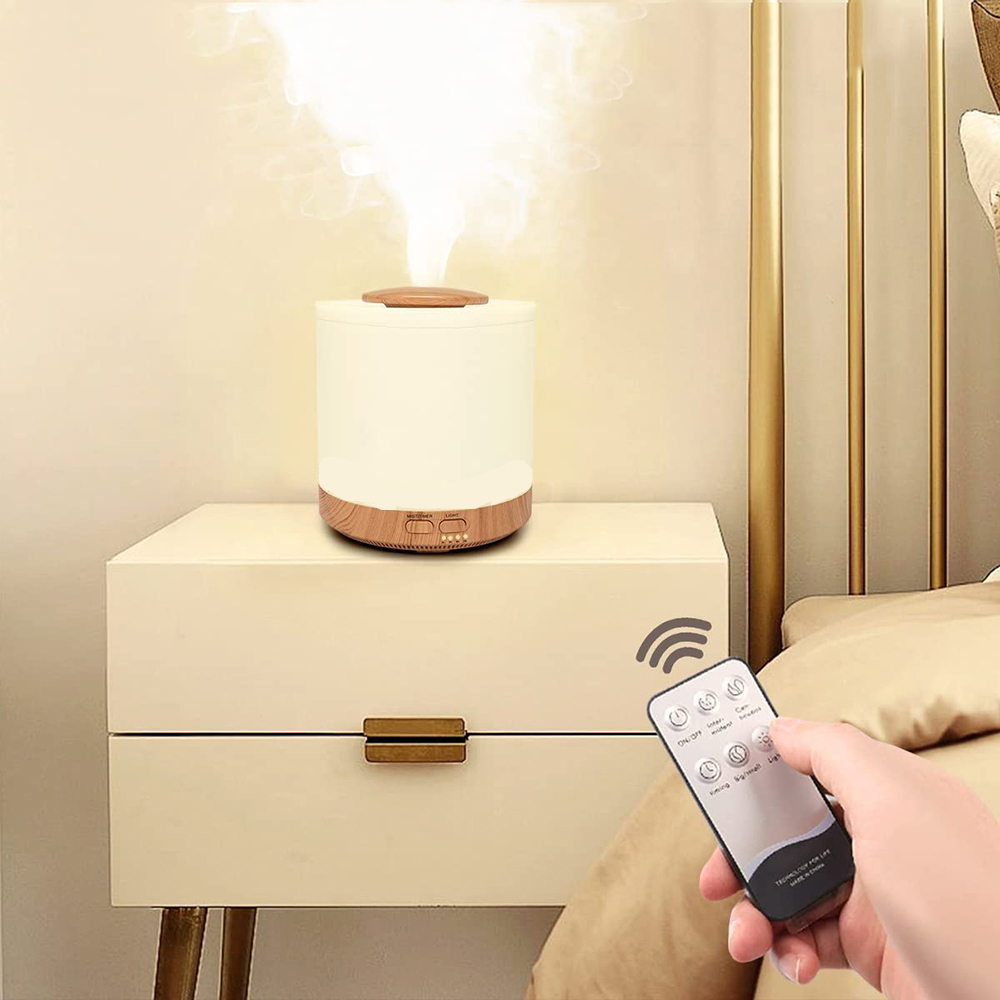 Auf einem Nachttisch steht ein weißer Diffusor mit Holzsockel. Eine Hand hält eine Fernbedienung. Rauch geht vom Diffusor aus. Im Hintergrund ist ein Bett.
