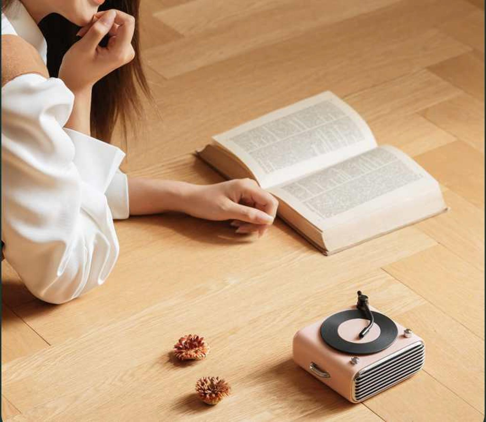 Eine Person ruht sich in der Nähe eines offenen Buches und eines rosa Plattenspielers auf einem Holzboden aus, mit zwei kleinen getrockneten Blumen.