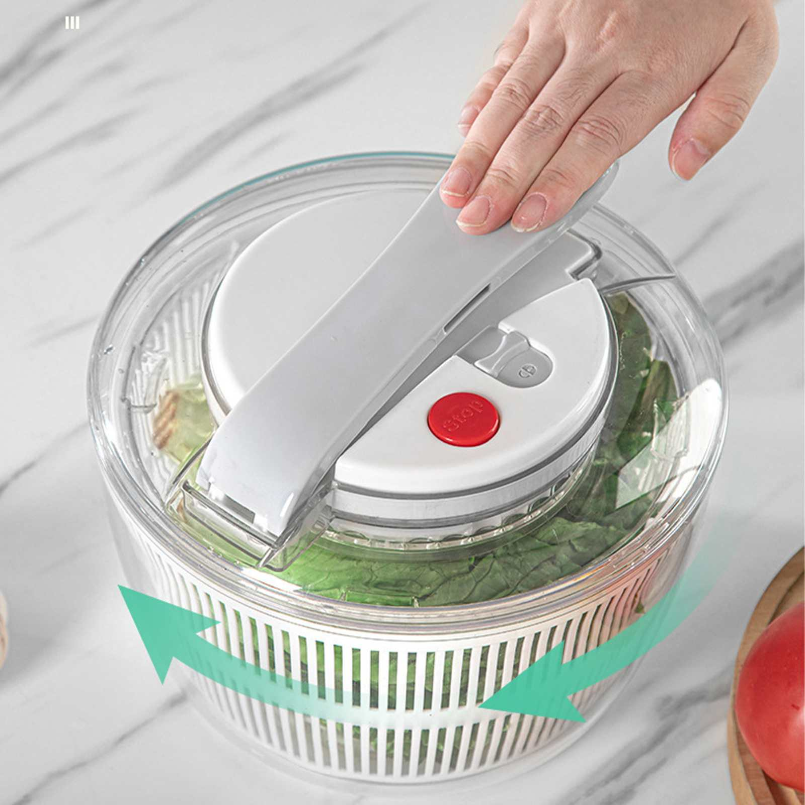 Eine Hand bedient eine Salatschleuder. Das Gerät ist transparent mit weißen Teilen, gefüllt mit grünem Salat und hat einen roten Stoppknopf.
